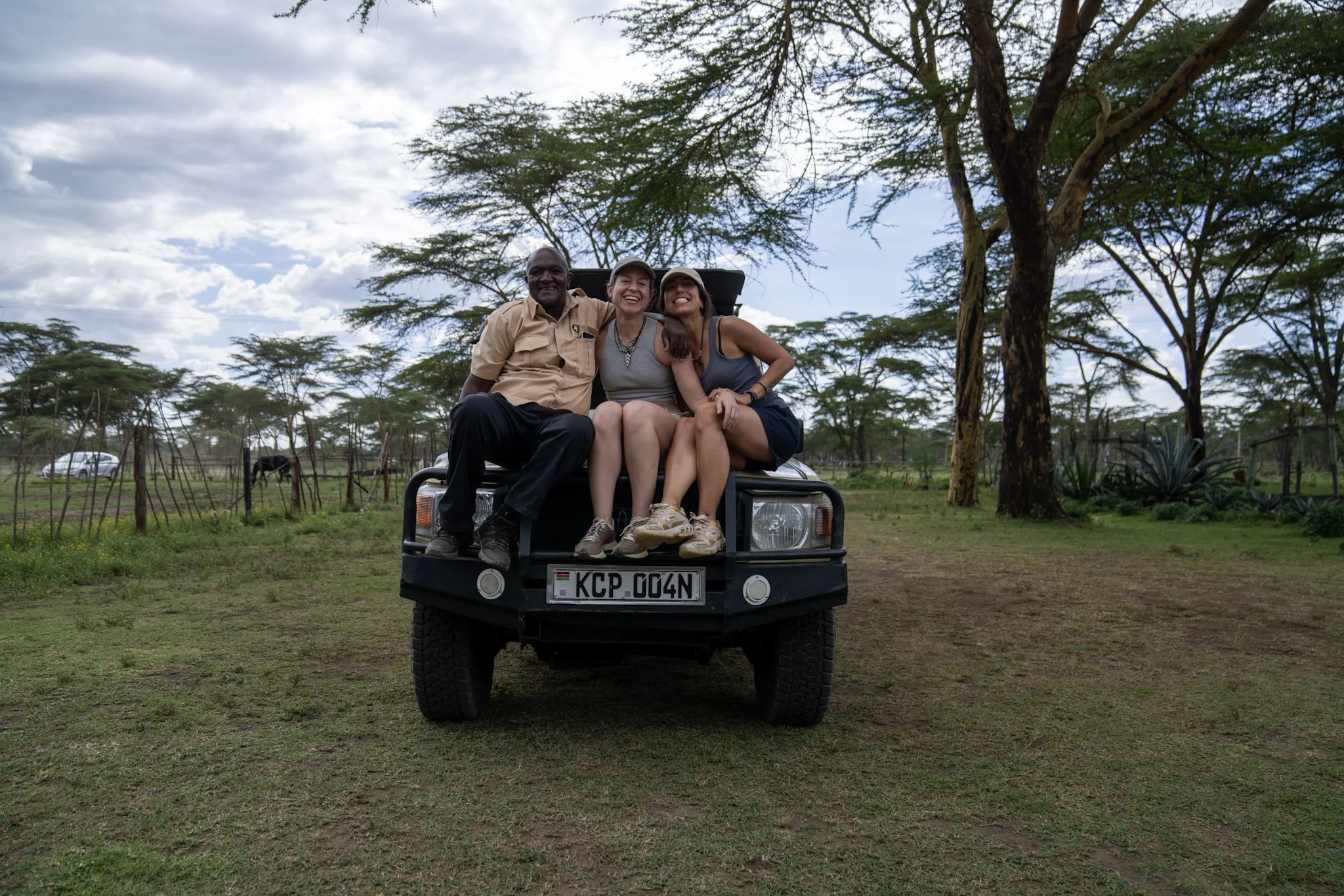 Three people sitting on the front of a safari vehicle in an open field with trees in the background.