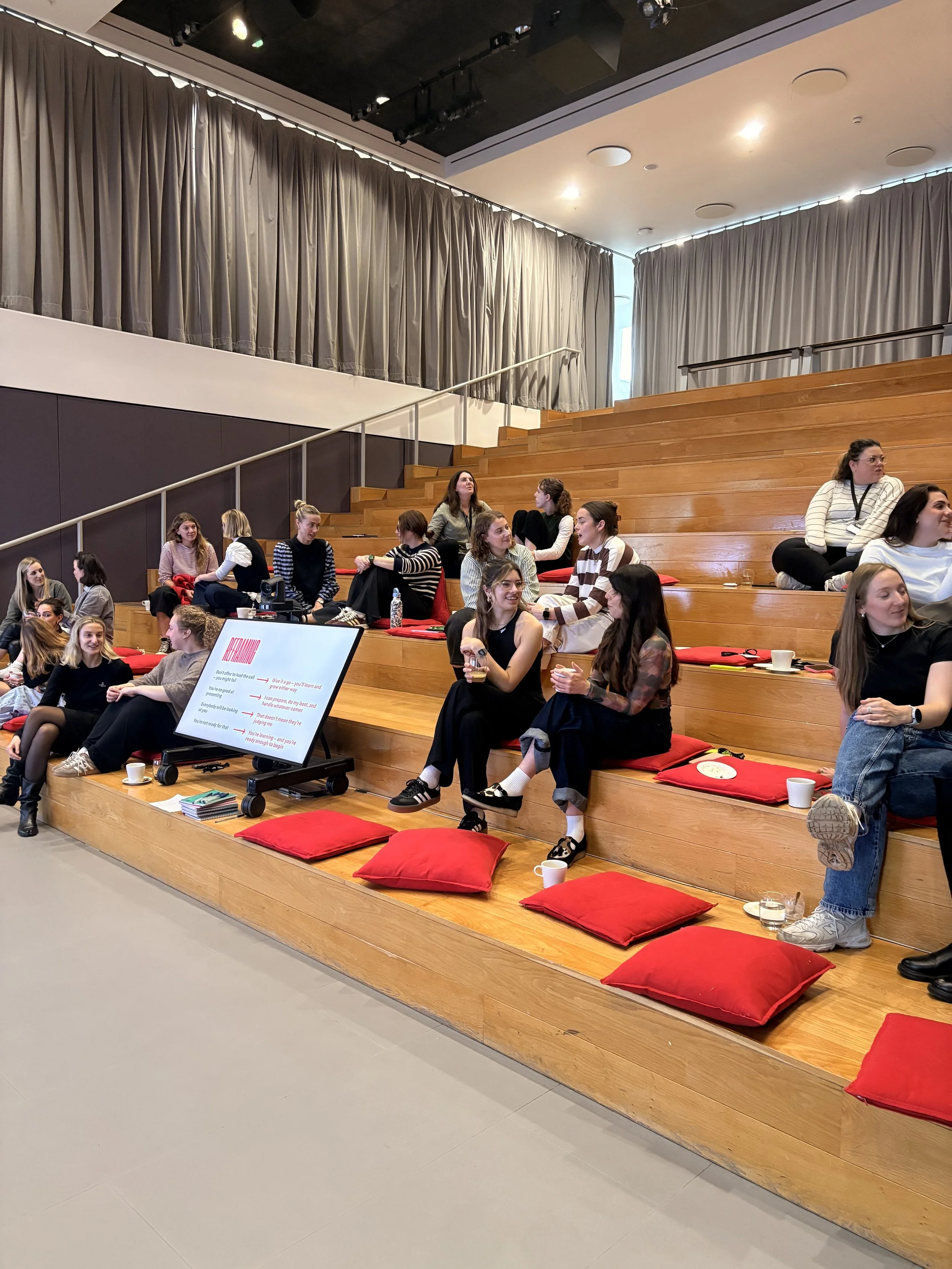 A group of women sitting on wooden tiered benches with red cushions in a modern conference or seminar room, some engaging in conversation, with a large digital screen displaying text on the floor in front of them.