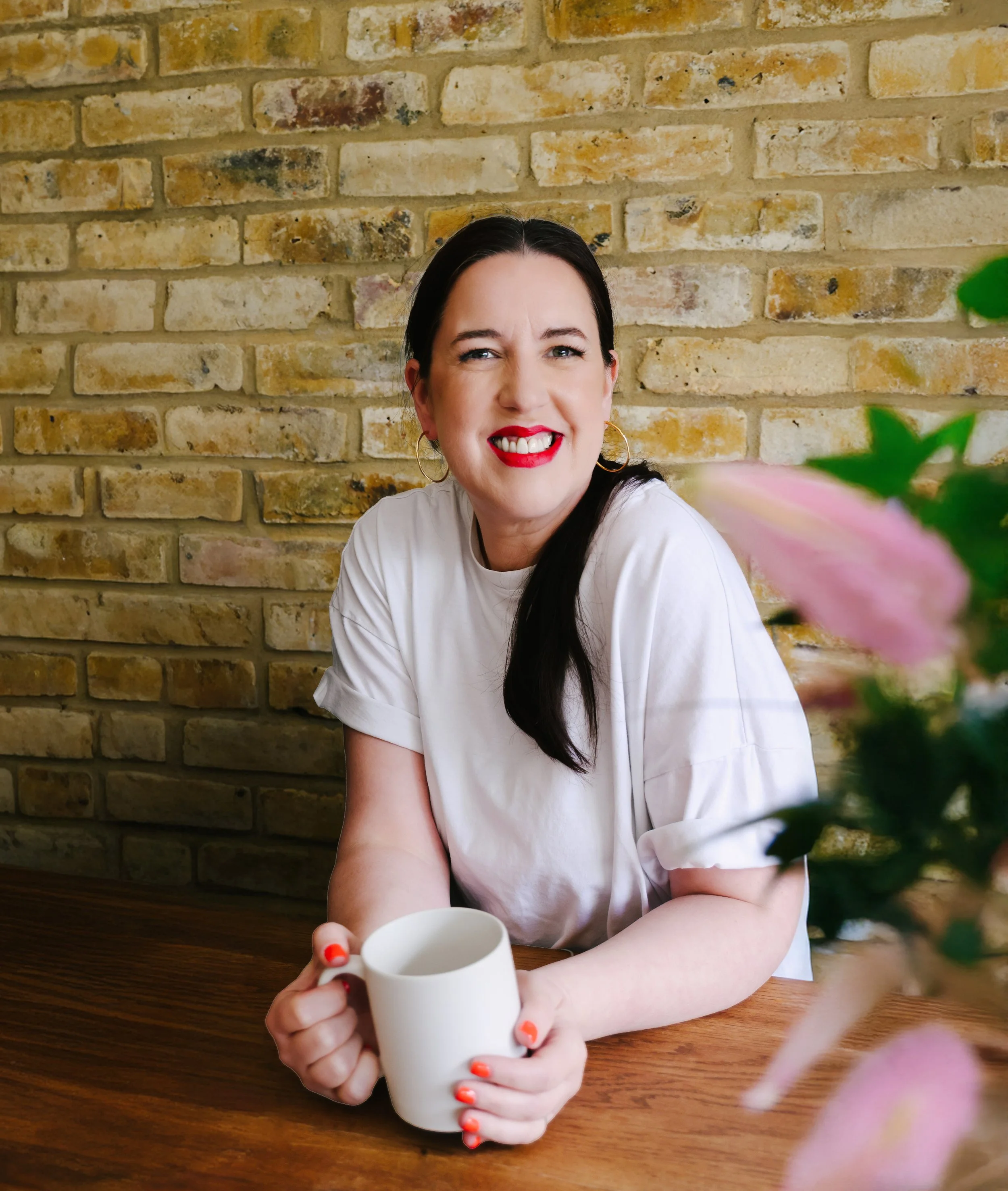 A woman with dark hair, wearing a white t-shirt and red lipstick, sitting at a wooden table in front of a brick wall, holding a white mug, with pink flowers and green leaves in the foreground.