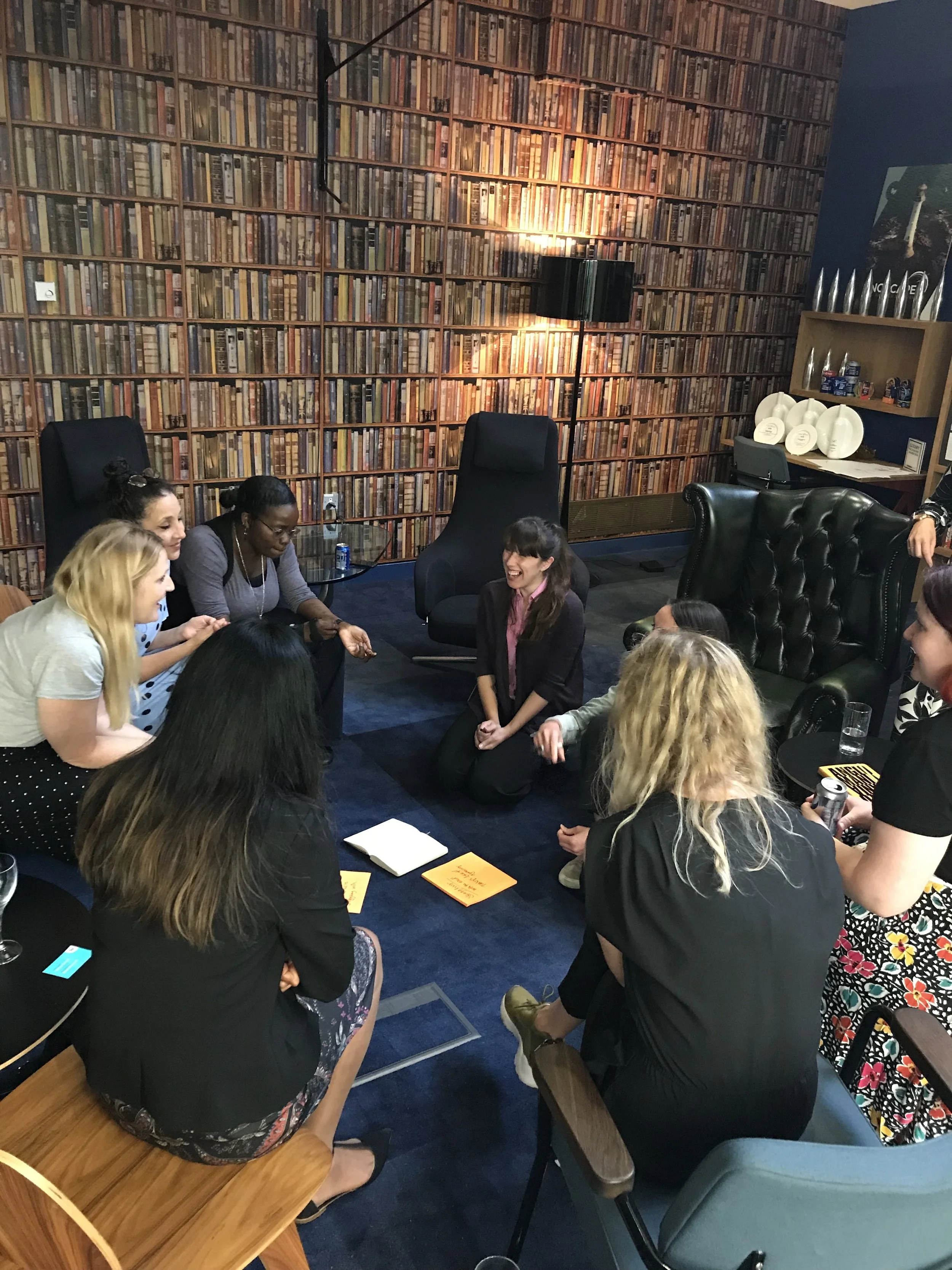 A group of women gathered in a circle in a cozy room with a bookshelf wallpaper, laughing and talking.