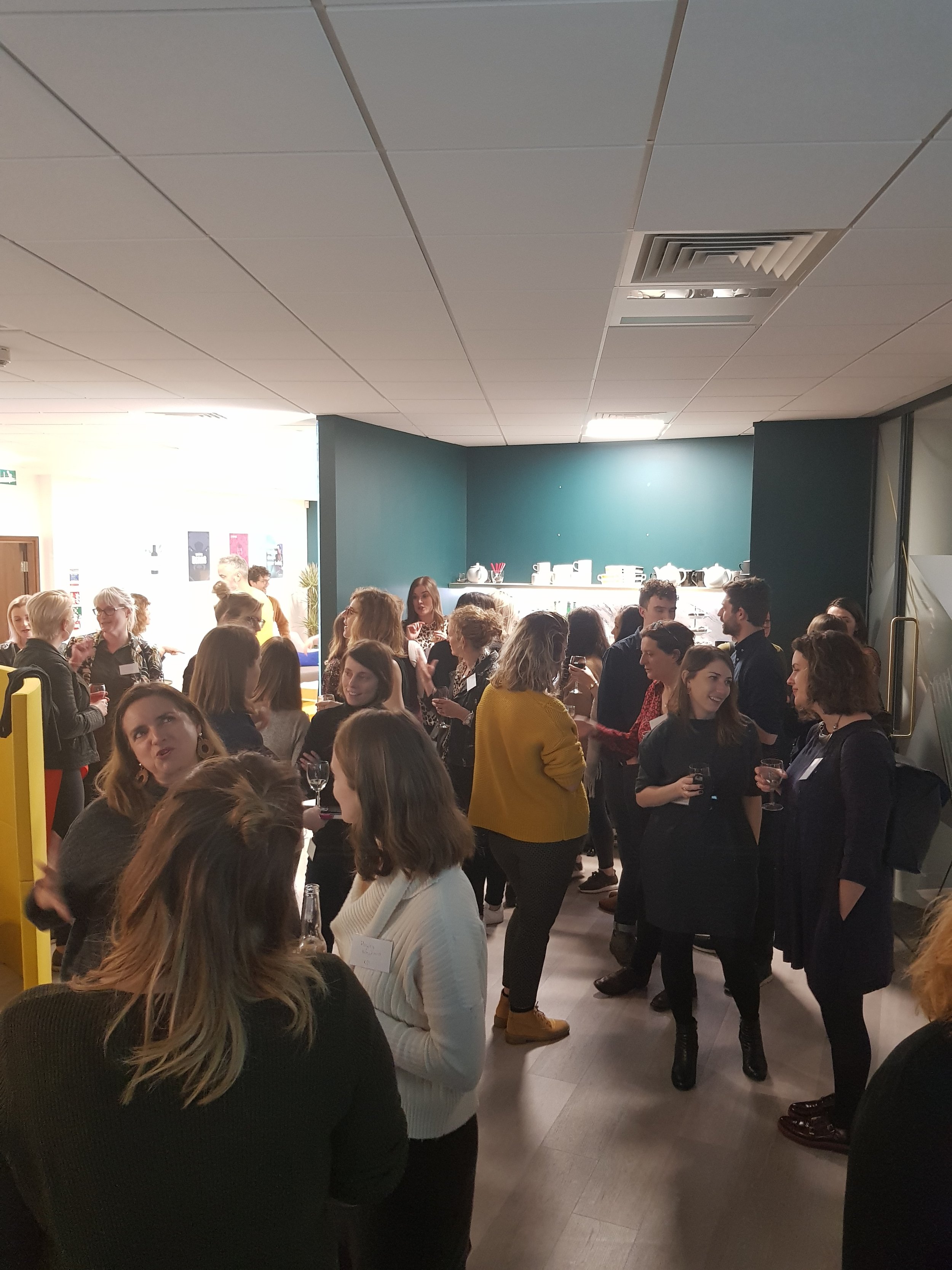 A social gathering of mostly women in an indoor venue, engaging in conversation, some holding glasses, with a building interior and a refreshment station in the background.