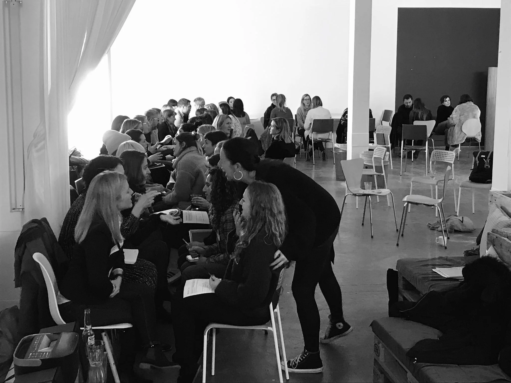 A group of people sitting in a room, engaging in conversation, with some holding papers and drinks, in a casual setting.