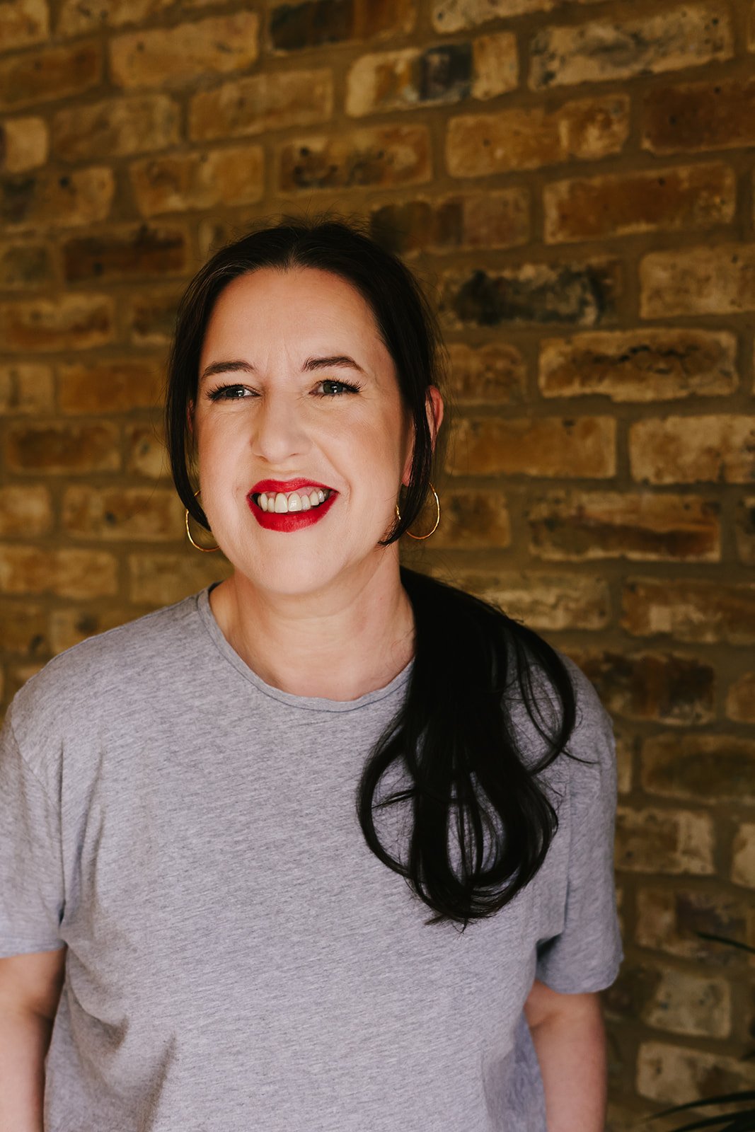 A woman with long black hair, wearing a gray t-shirt and gold hoop earrings, smiling in front of a brick wall.