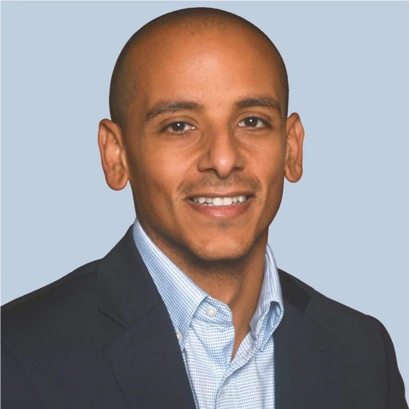 Headshot of a man with a shaved head wearing a dark blazer over a light blue collared shirt, smiling against a light blue background.