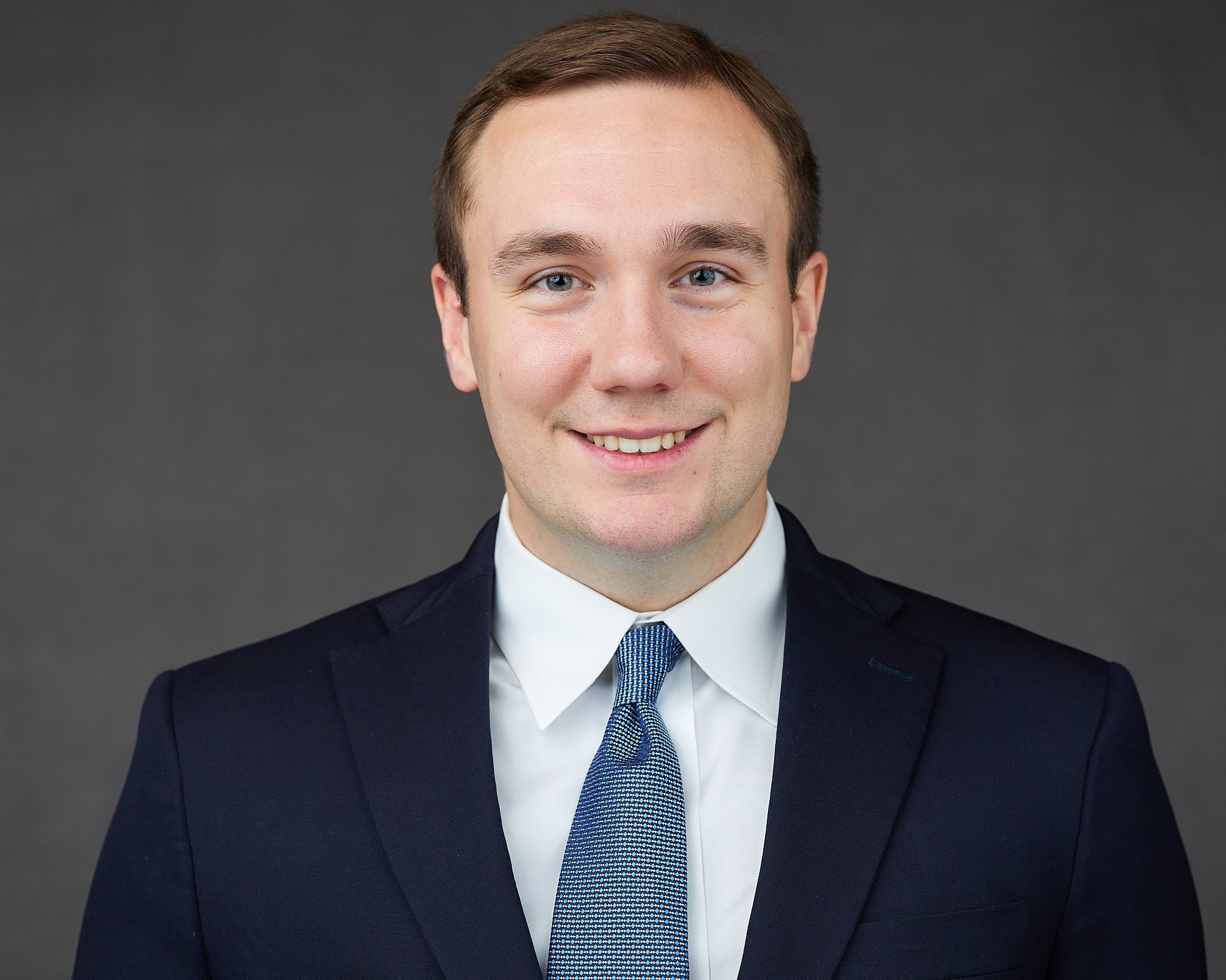 A young man with light skin, wearing a dark suit, white shirt, and blue patterned tie, smiling at the camera.