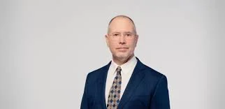 Portrait of a man in a navy suit, white shirt, and patterned tie, against a plain background.