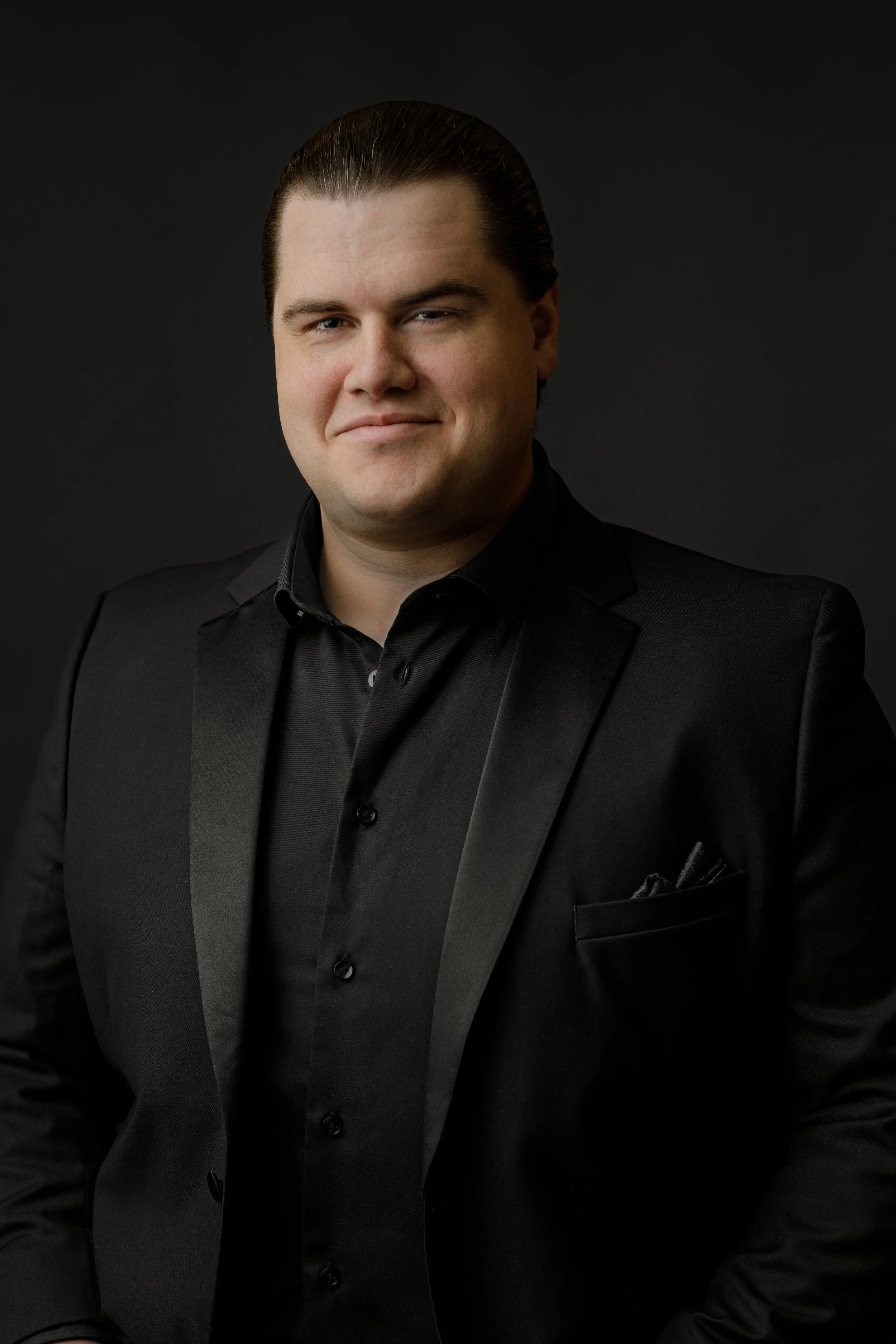 A man with slicked-back dark hair, wearing a black suit and shirt, posing against a dark background.