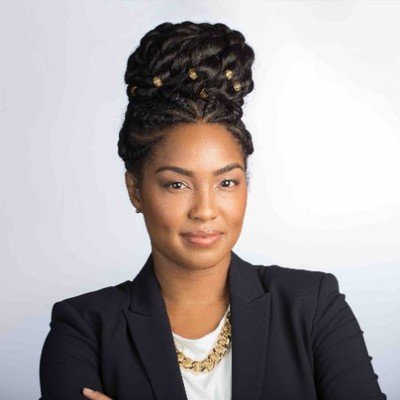 Portrait of a woman with braided hair styled in an updo, wearing a black blazer and a gold chain necklace, against a plain light background.