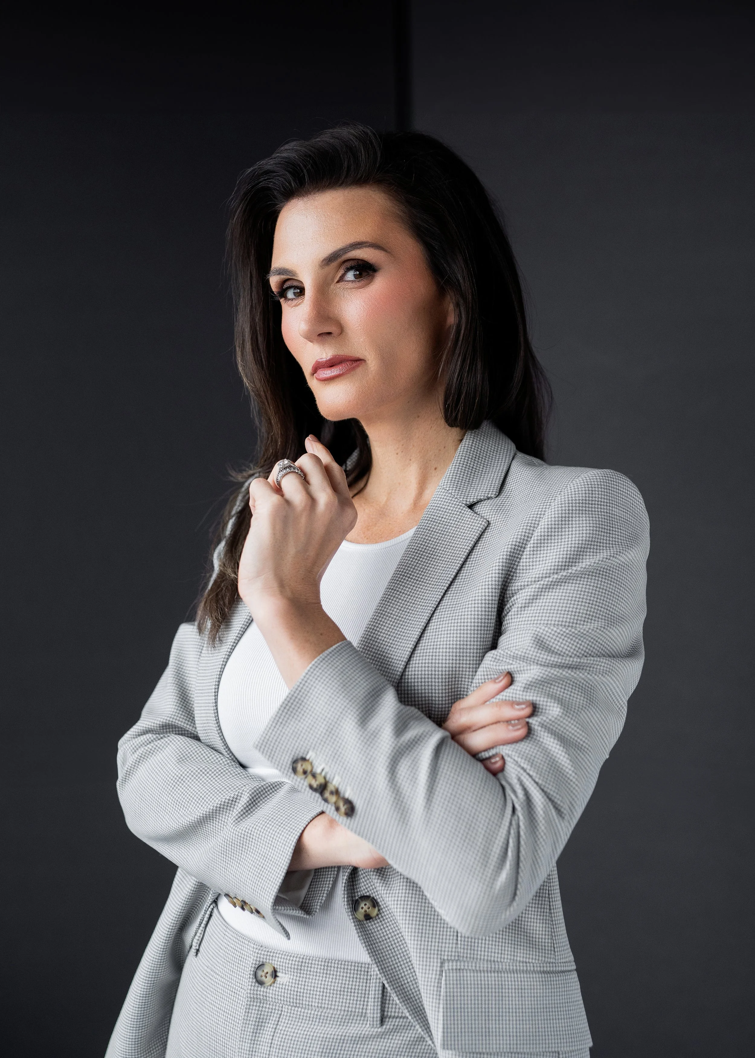 A confident woman with dark hair and fair skin wearing a light gray blazer and white top, posing with her arms crossed and looking at the camera against a dark background.