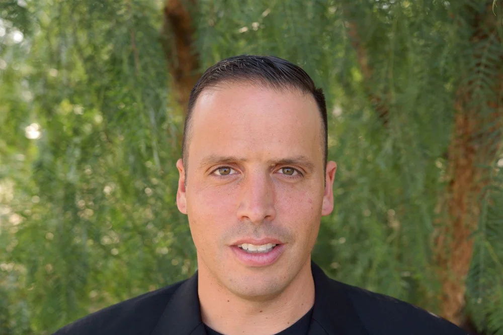 A man with short dark hair and light skin, wearing a black jacket, standing outdoors in front of green foliage.
