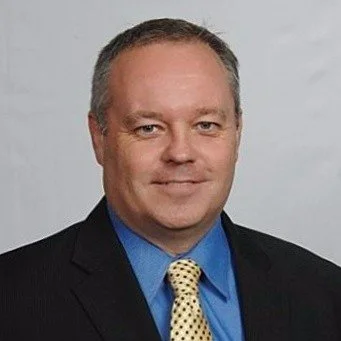 Professional headshot of a man in a suit with a blue shirt and yellow patterned tie, against a gray background.