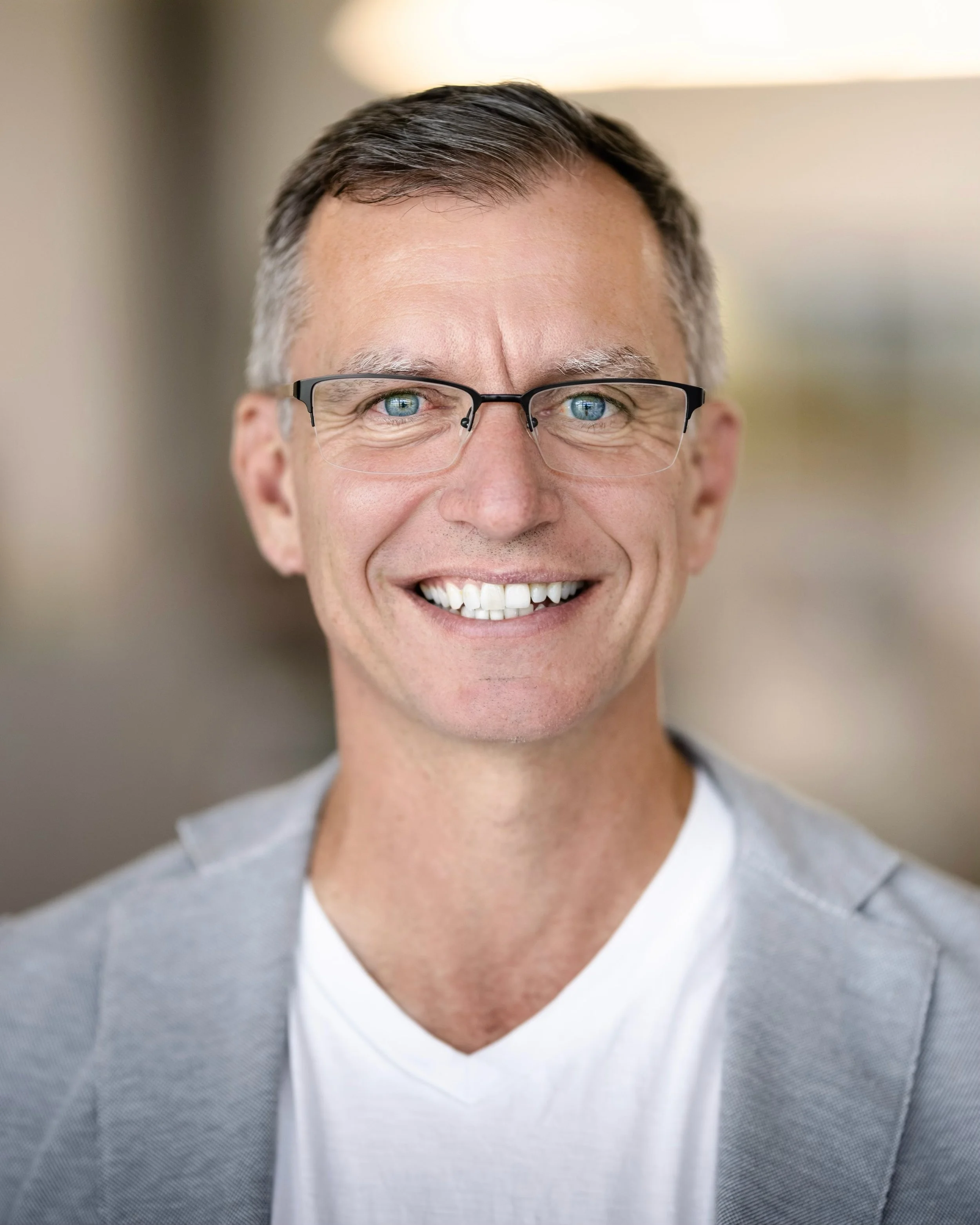 Portrait of a middle-aged man with glasses, smiling, wearing a light gray blazer and white T-shirt, in an indoor setting with blurred background.
