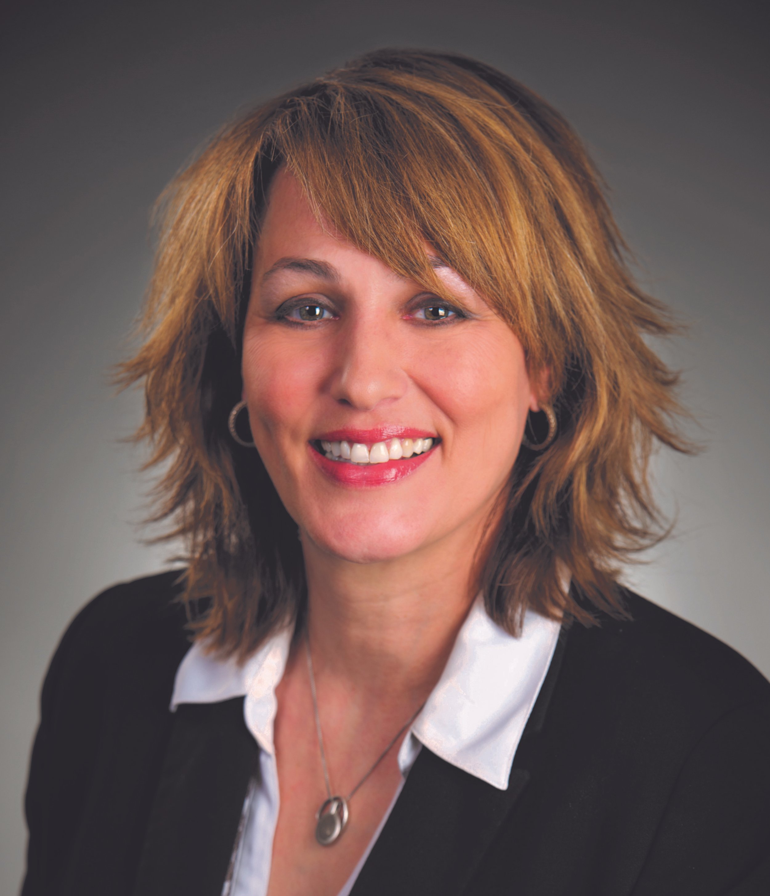 A woman with short, layered, light brown hair smiling at the camera, wearing a black blazer over a white shirt with a silver necklace and hoop earrings.