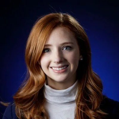 Portrait of a young woman with red hair, smiling, wearing a white turtleneck and a dark blazer against a dark blue background.