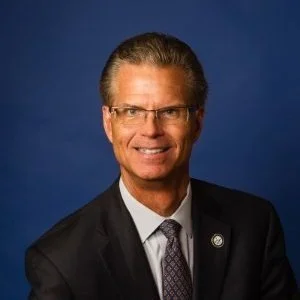 A professional man in a suit and tie, wearing glasses, smiling against a dark blue background.