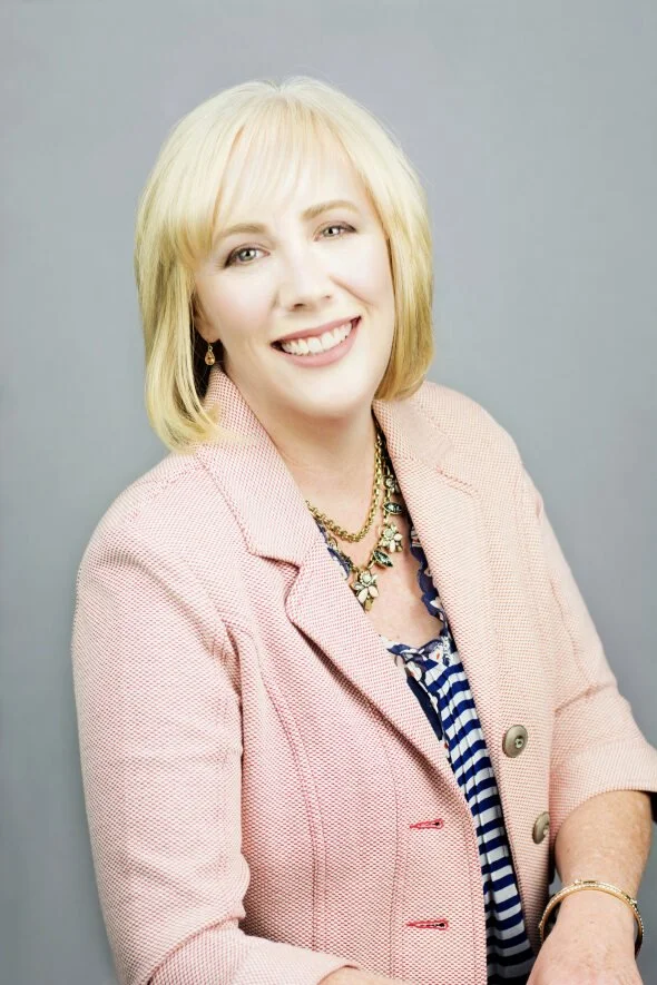 A woman with blonde hair smiling, wearing a pink blazer over a blue and white striped top, accessorized with gold jewelry, against a gray background.