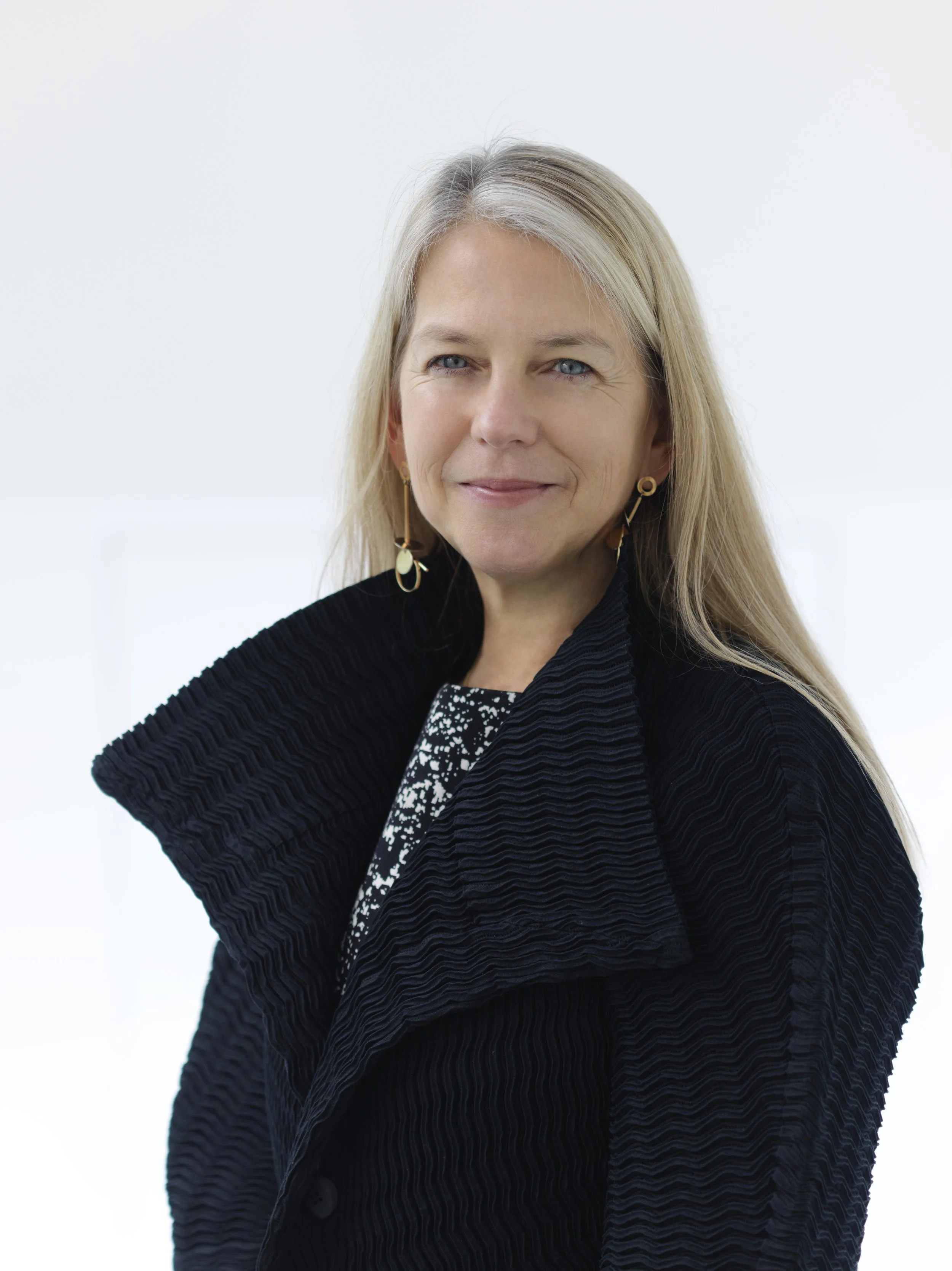 A blonde woman wearing a black textured coat and patterned top, smiling softly against a plain white background.