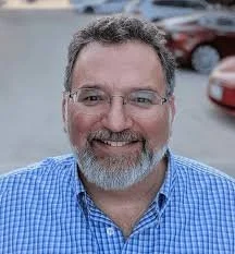 A smiling man with glasses and a beard, wearing a blue checkered shirt, outdoors with cars in the background.