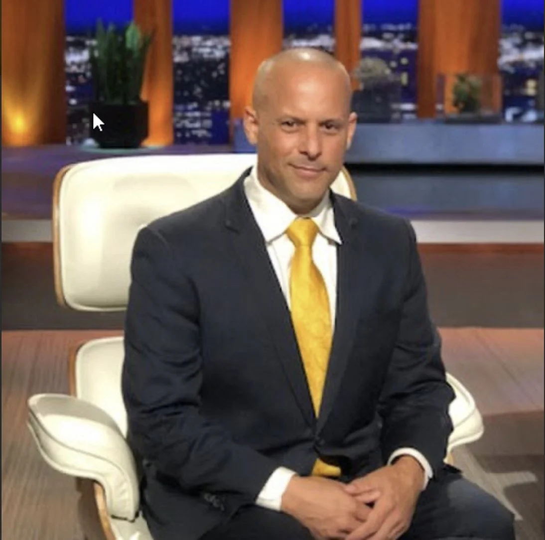 A bald man wearing a dark suit, white shirt, and yellow tie sitting in a modern white chair on a television talk show set with a cityscape background at night.