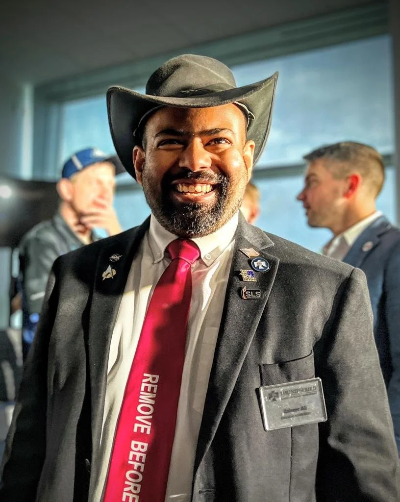 A smiling man wearing a cowboy hat, suit, red tie with white text, and a badge, at a professional or conference event with other people in the background.