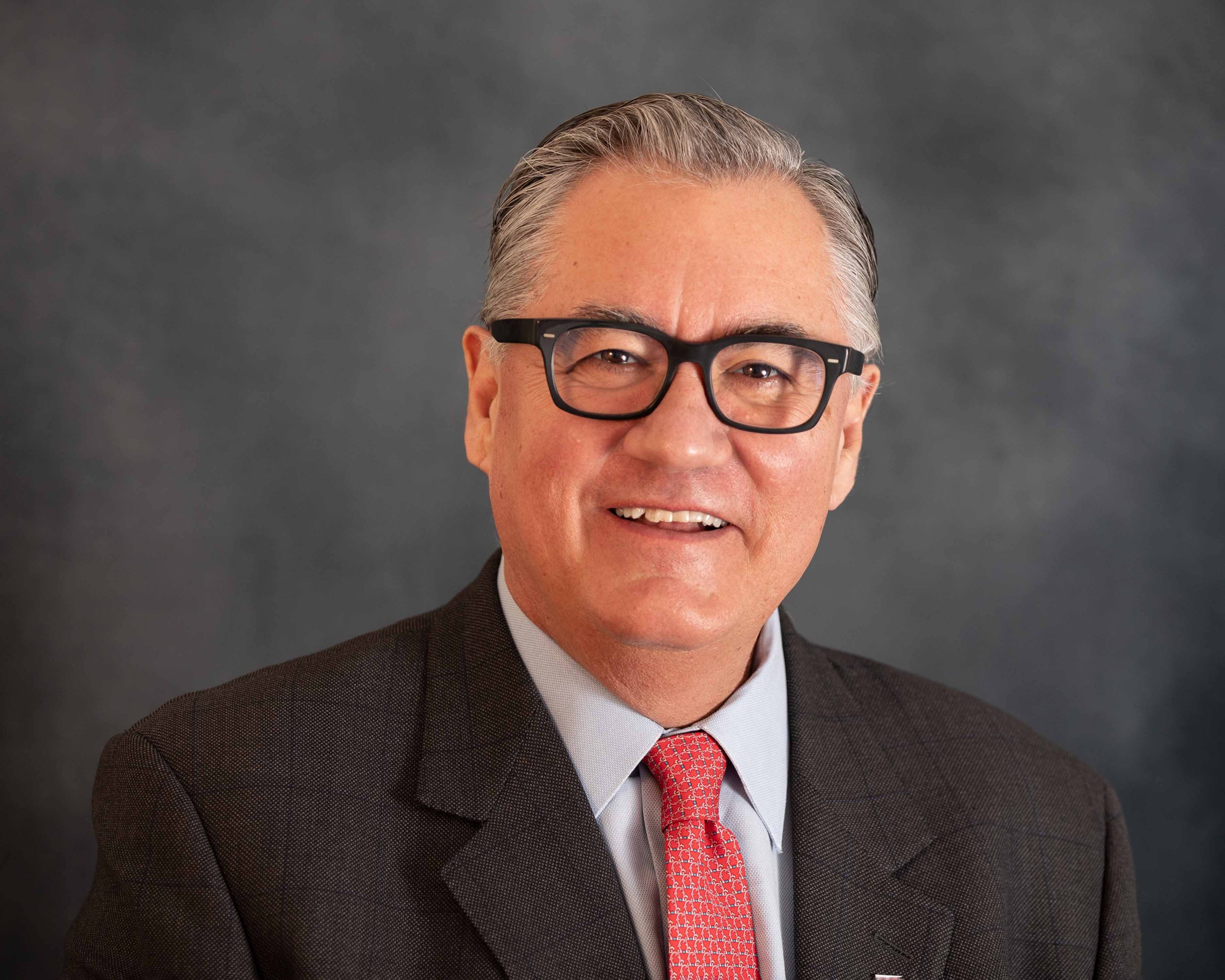 Portrait of a smiling man with gray hair, wearing glasses, a dark suit, white shirt, and red tie, against a gray background.