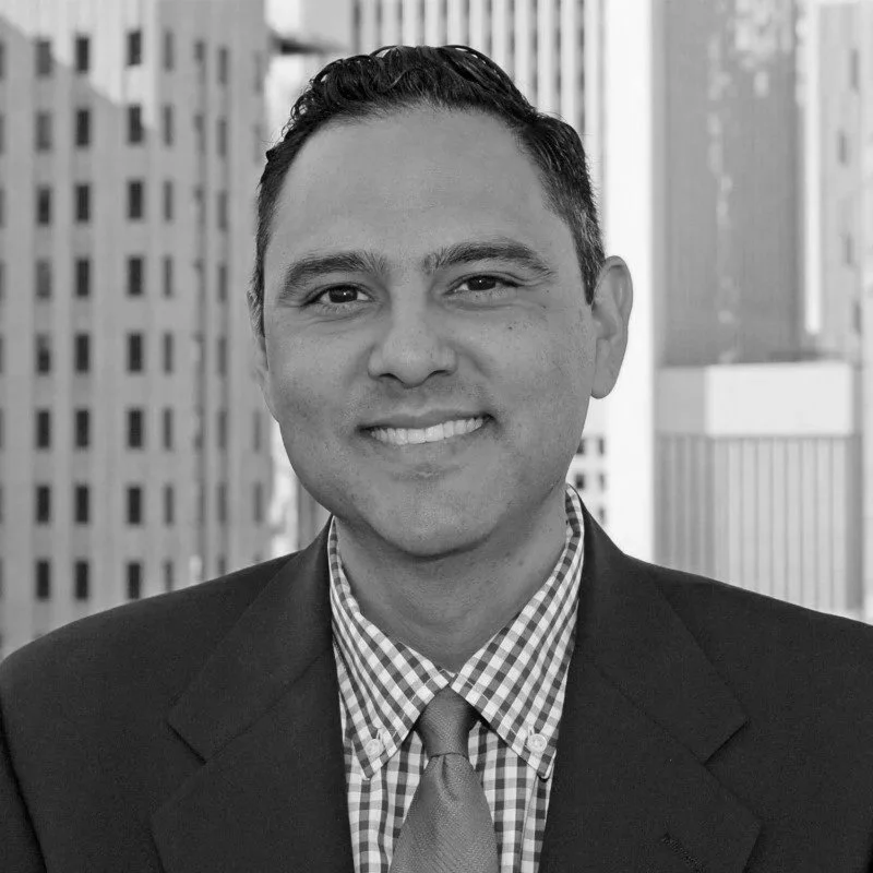 A smiling man in a business suit with a checkered shirt and tie, standing outdoors against a backdrop of tall city buildings.