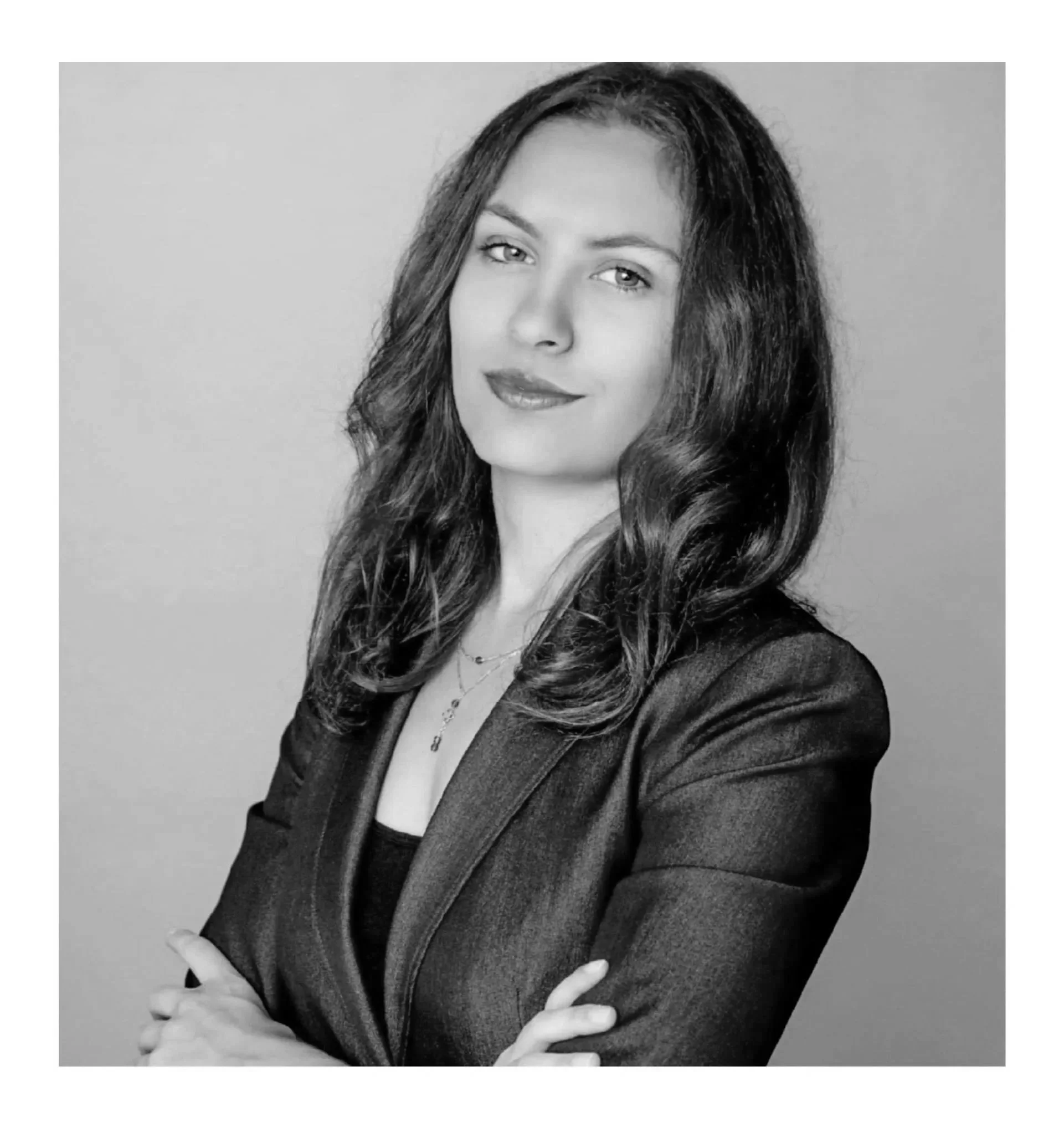 A woman with shoulder-length brown hair and red lipstick, standing against a neutral background, wearing a dark blazer and necklace, with her arms crossed.