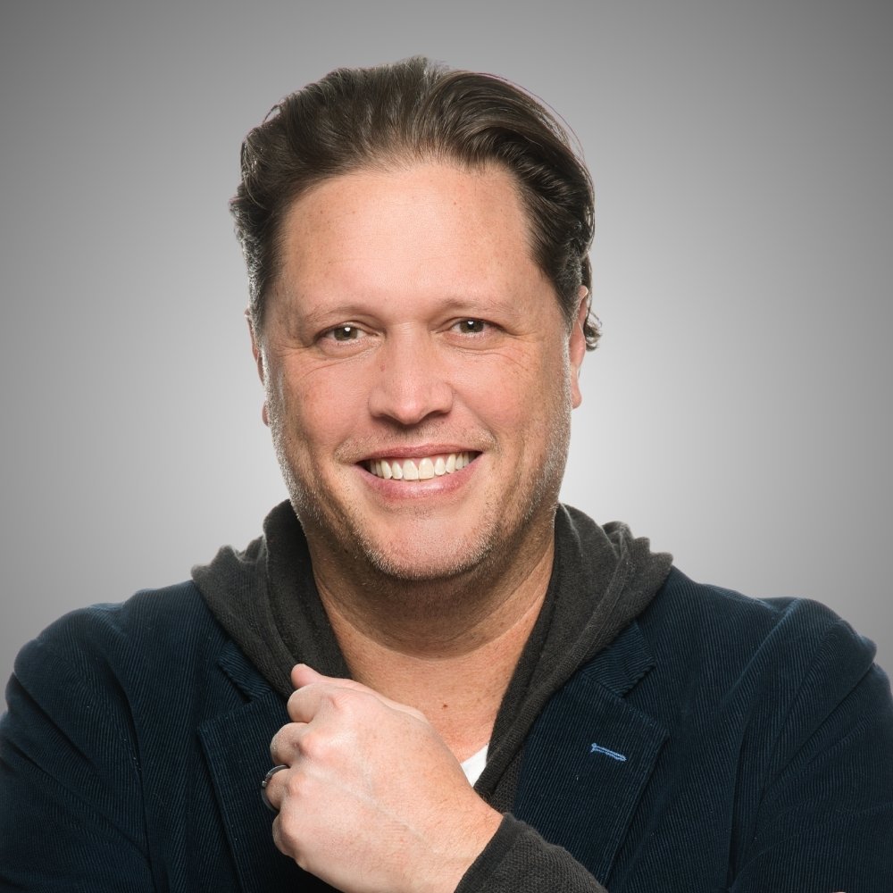 Portrait of a smiling man with brown hair, wearing a dark jacket and hoodie, against a light grey background.