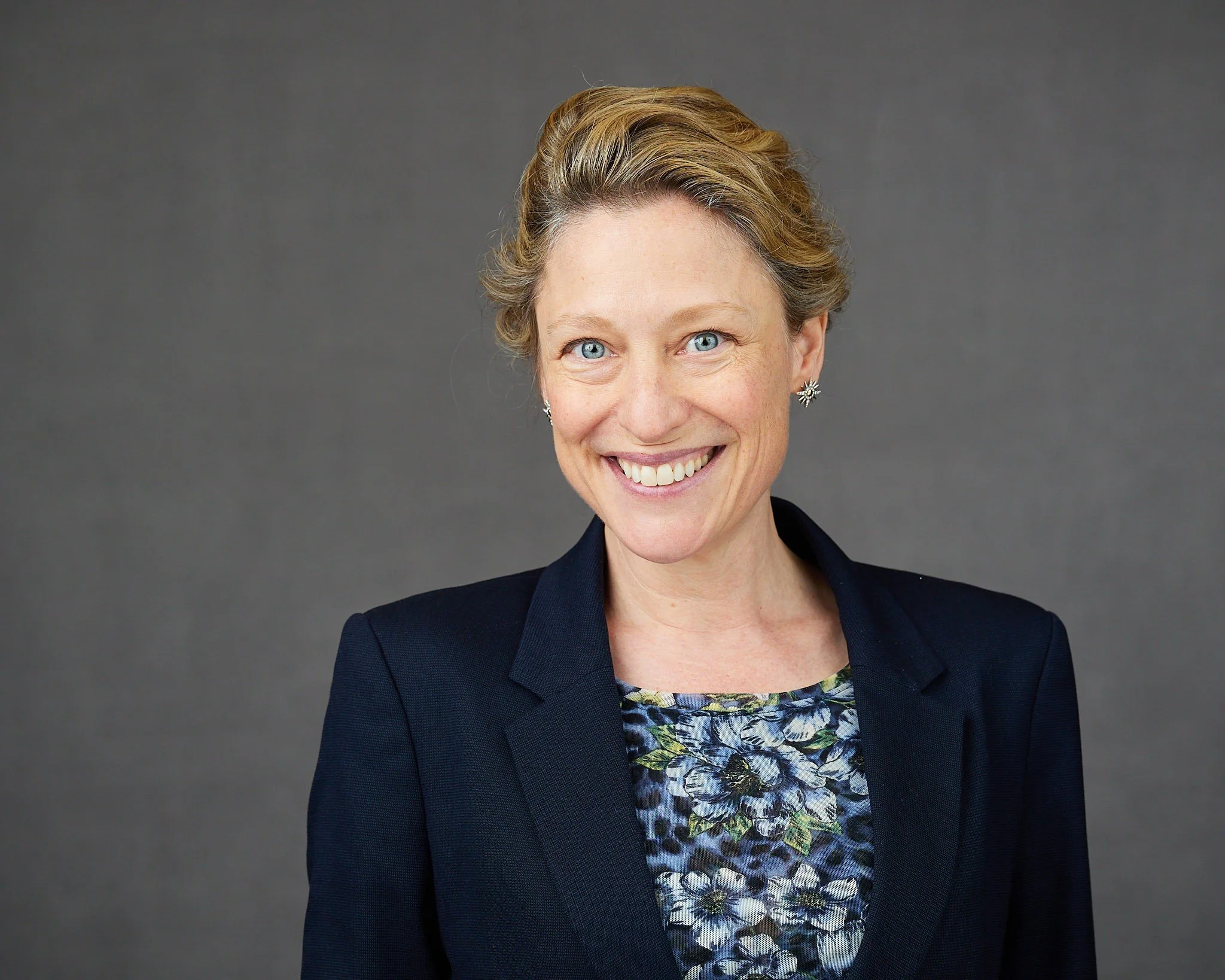 A professional woman with short blonde hair and blue eyes smiling, wearing a dark blazer over a floral blouse, standing against a plain gray background.