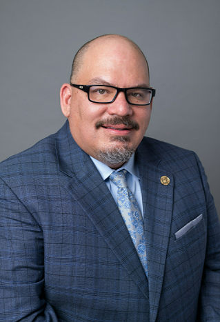 Portrait of a bald man with glasses wearing a blue checkered suit and a light blue patterned tie against a gray background.