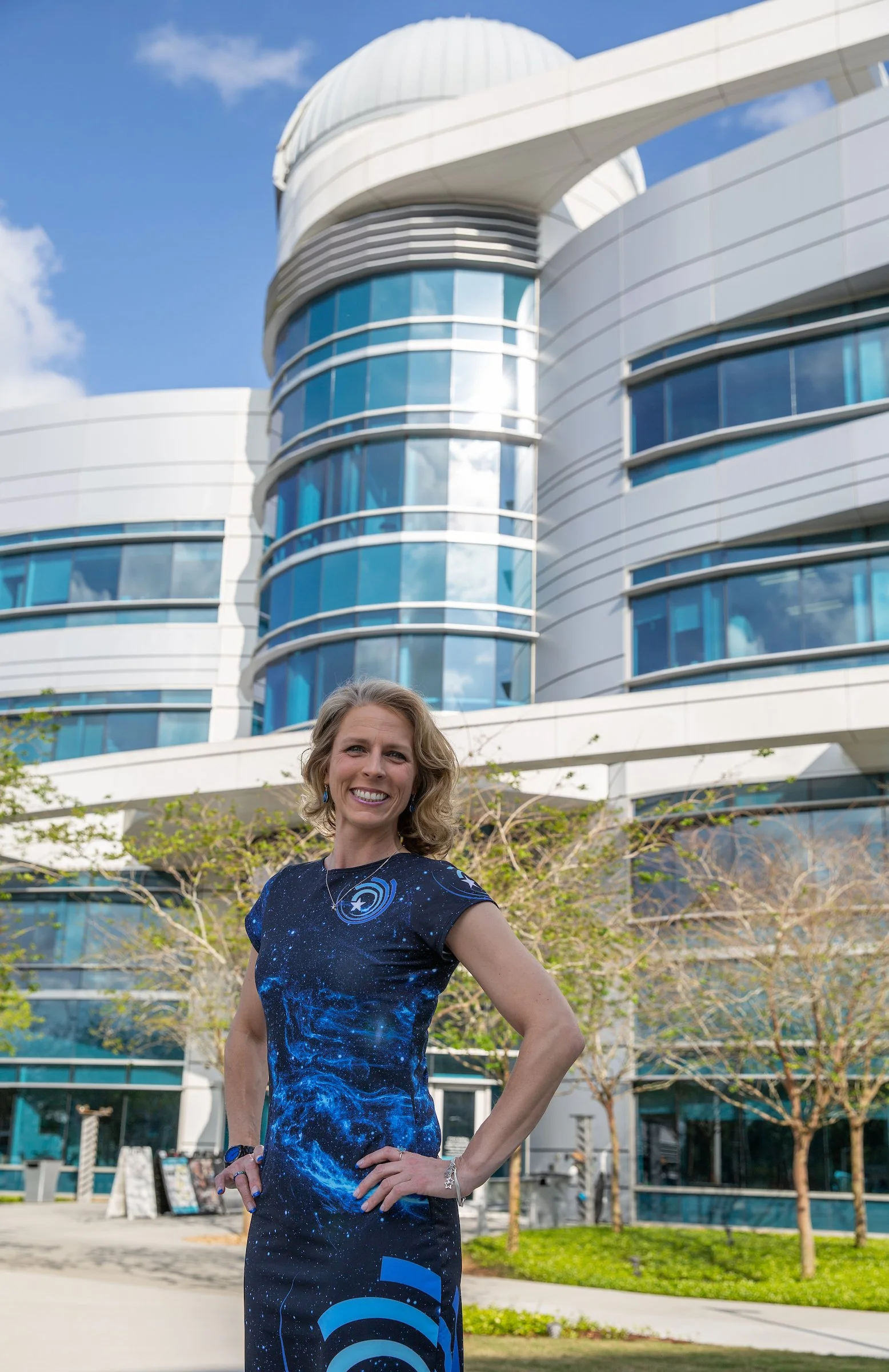 A woman in a blue space-themed dress standing outdoors in front of a modern building with curved glass and white exterior, smiling at the camera.