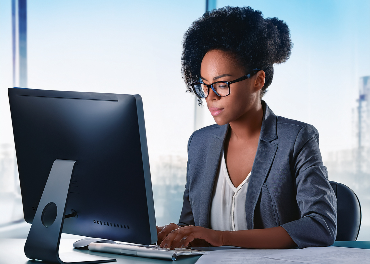 A woman with curly black hair and glasses working at a computer in an office with large windows.
