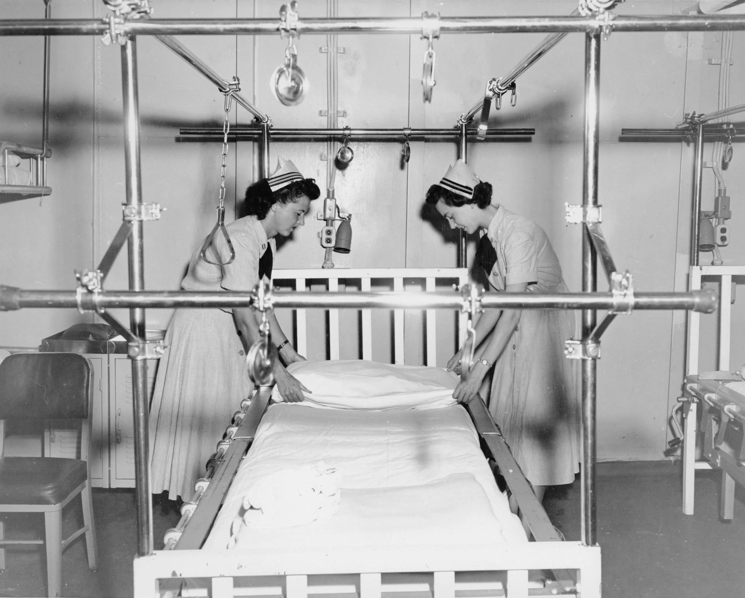 Black-and-white historical photo of nurses preparing a patient bed in an early hospital ward.