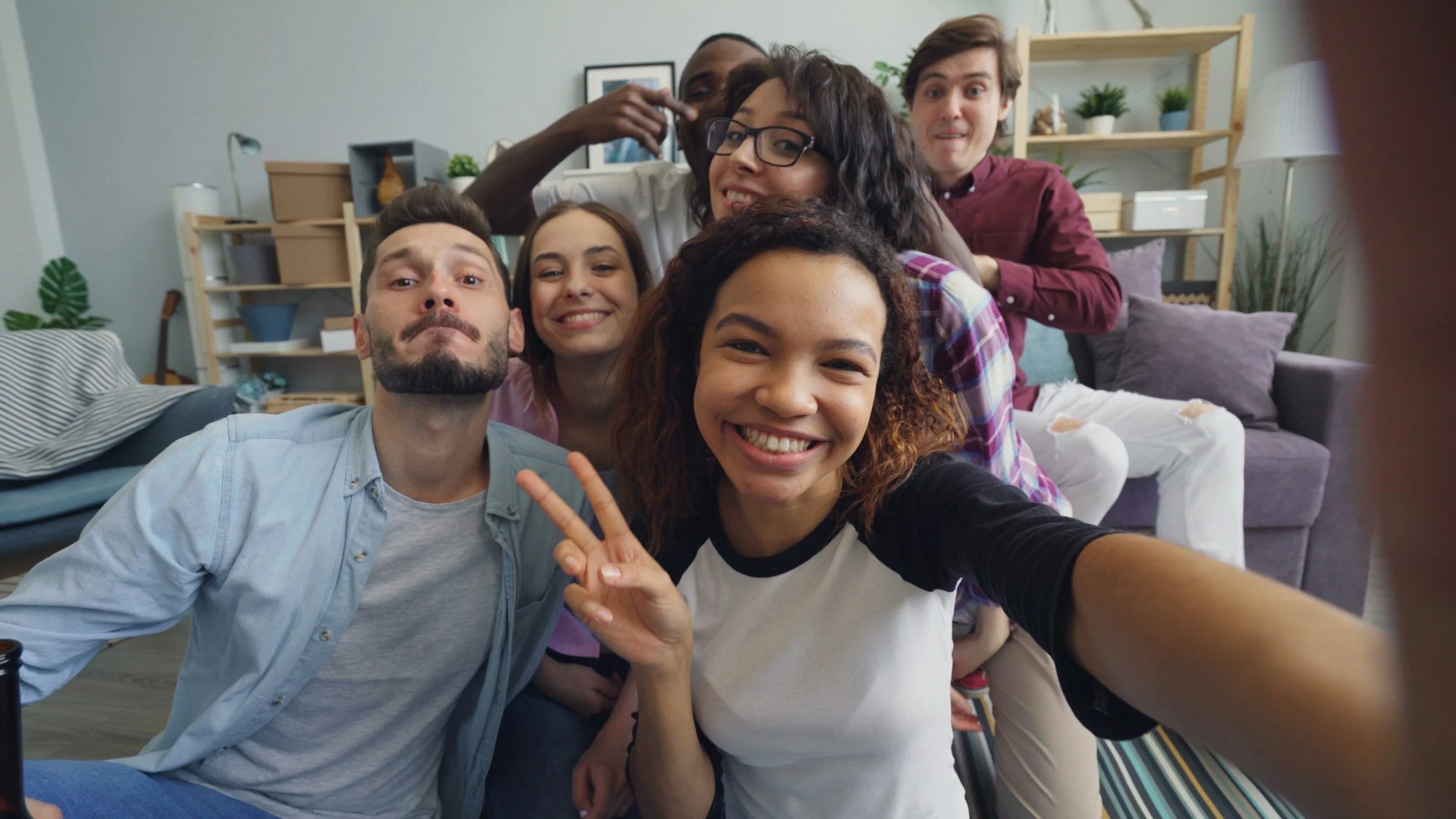 Group of six diverse friends smiling and taking a selfie together in a living room.