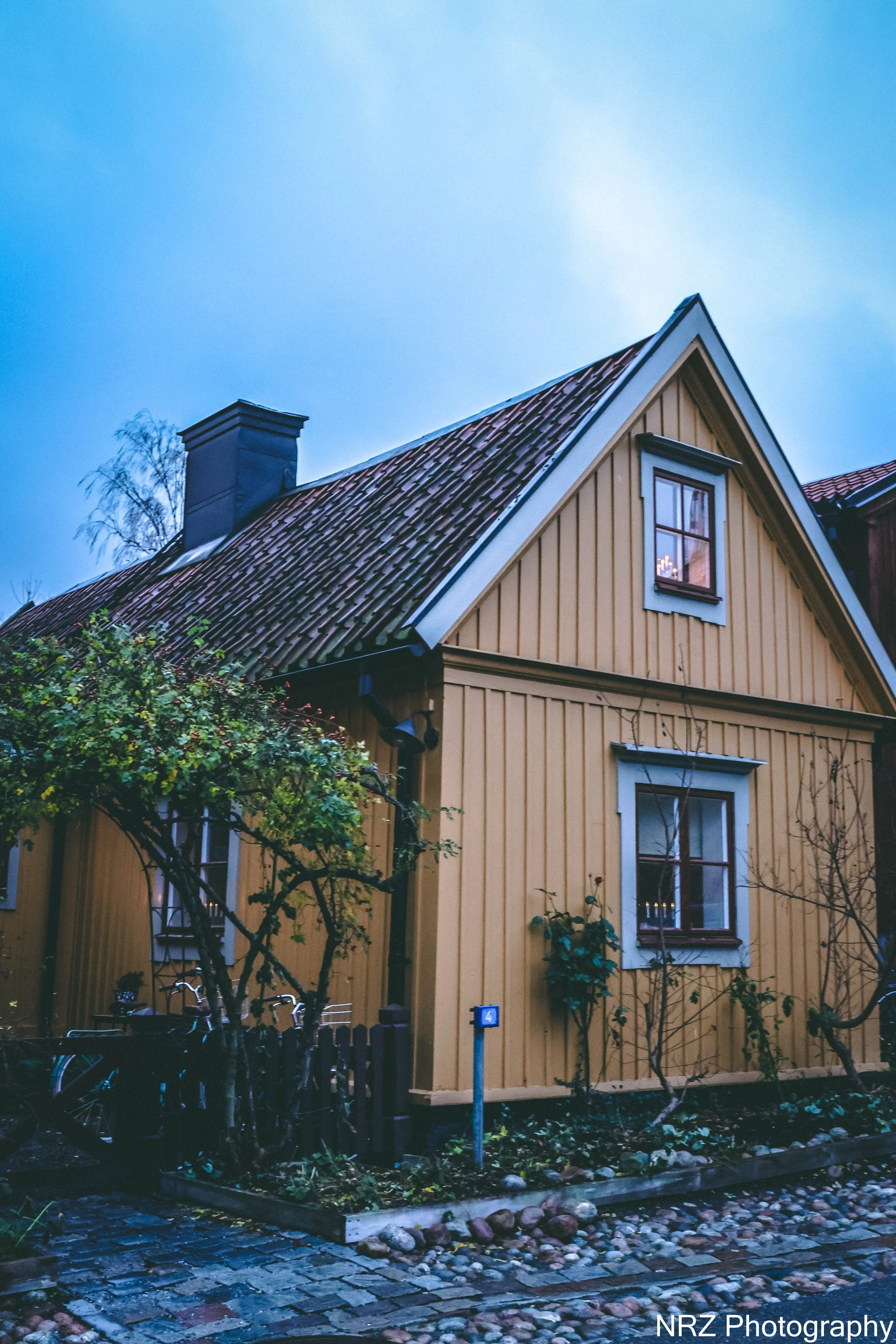 "Vinyl och vinterljus i hus nummer fyra"
Det gula huset med den branta taknocken låg tyst under snöns mjuka filt. Ljuset från fönstren fladdrade som levande minnen, och inne i stugan sprakade den öppna spisen med en glöd som luktade kåda och trygghet