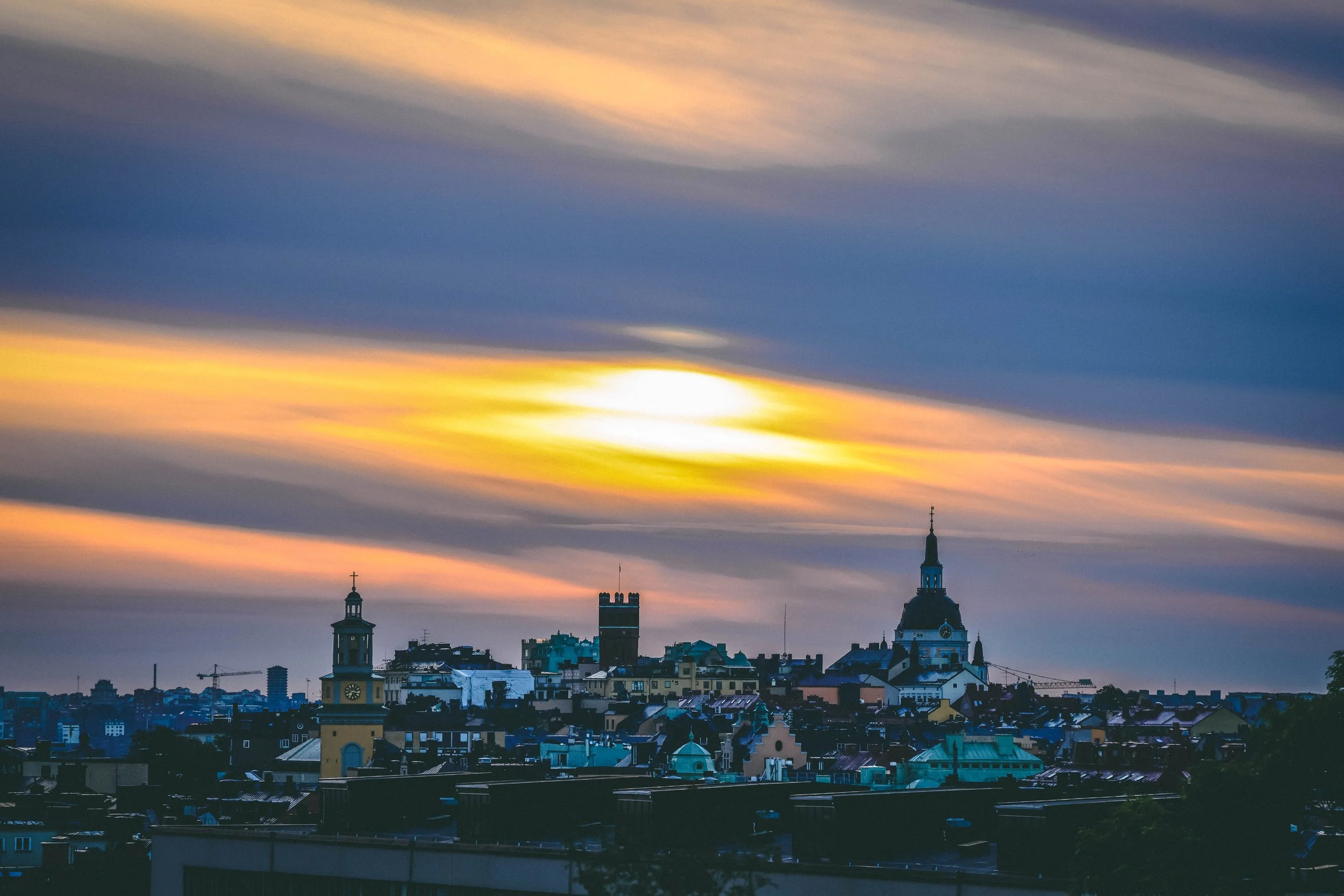 Golden breath over a waking city.
The first light of autumn spills across the rooftops, casting long shadows and warming the cool air with a soft amber glow. Church spires and towers stretch toward the sky, silhouetted against streaks of morning fir