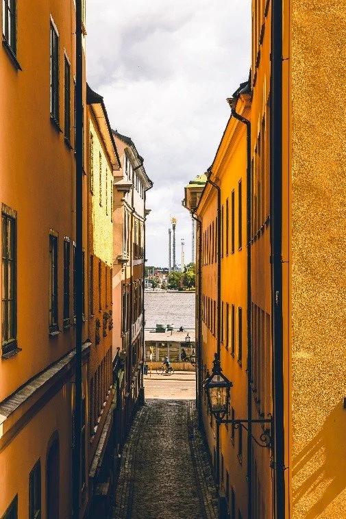 The cobblestone alley glowed in the late afternoon light, warm as a fresh croissant. The buildingsâpainted in shades of marmalade and mustardâleaned in like nosy aunties, watching the drama unfold below. Across the water, the amusement park spun and