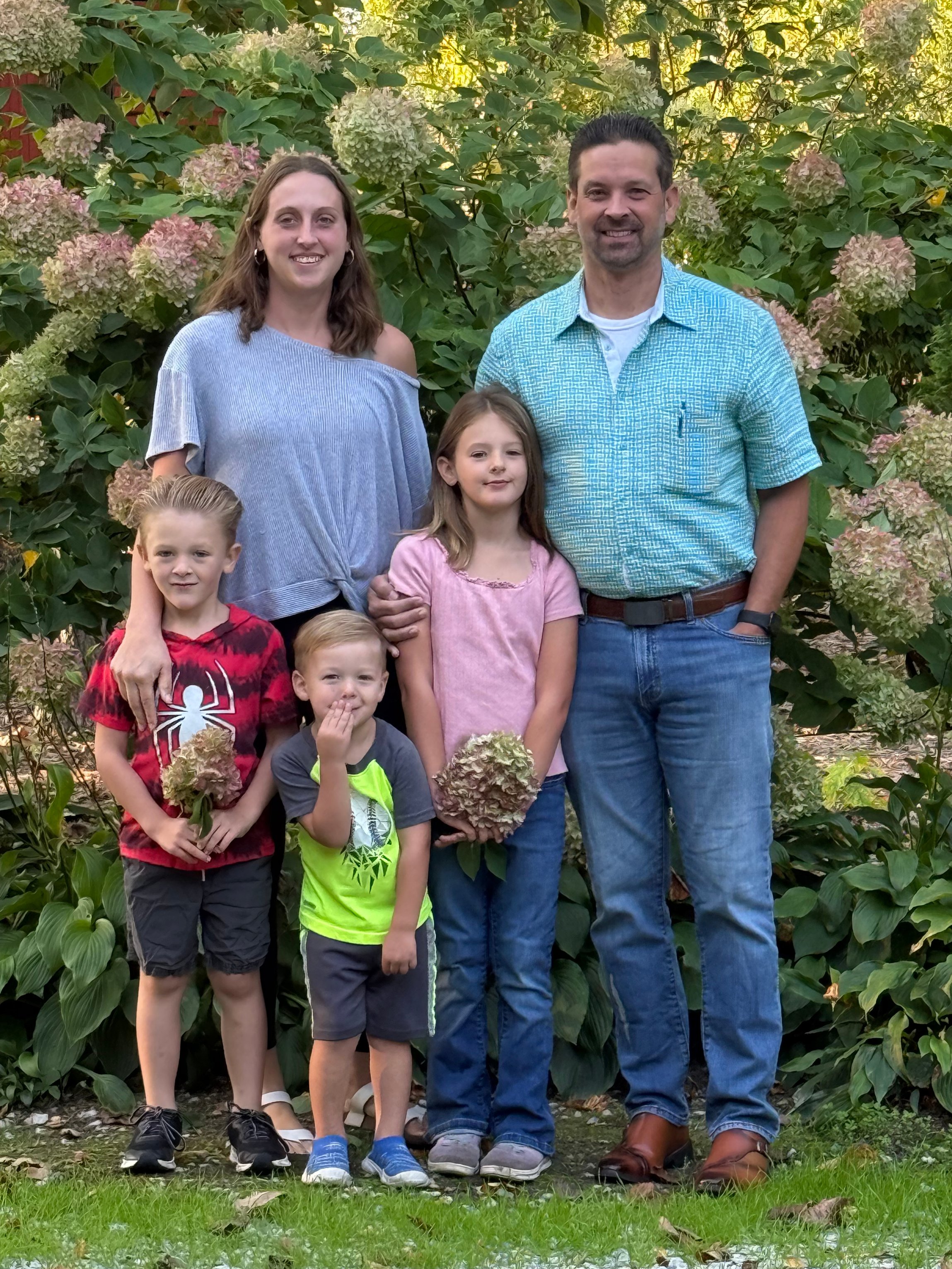 Family outdoor portrait in front of flowering shrub, including two adults and three children, some holding flowers.