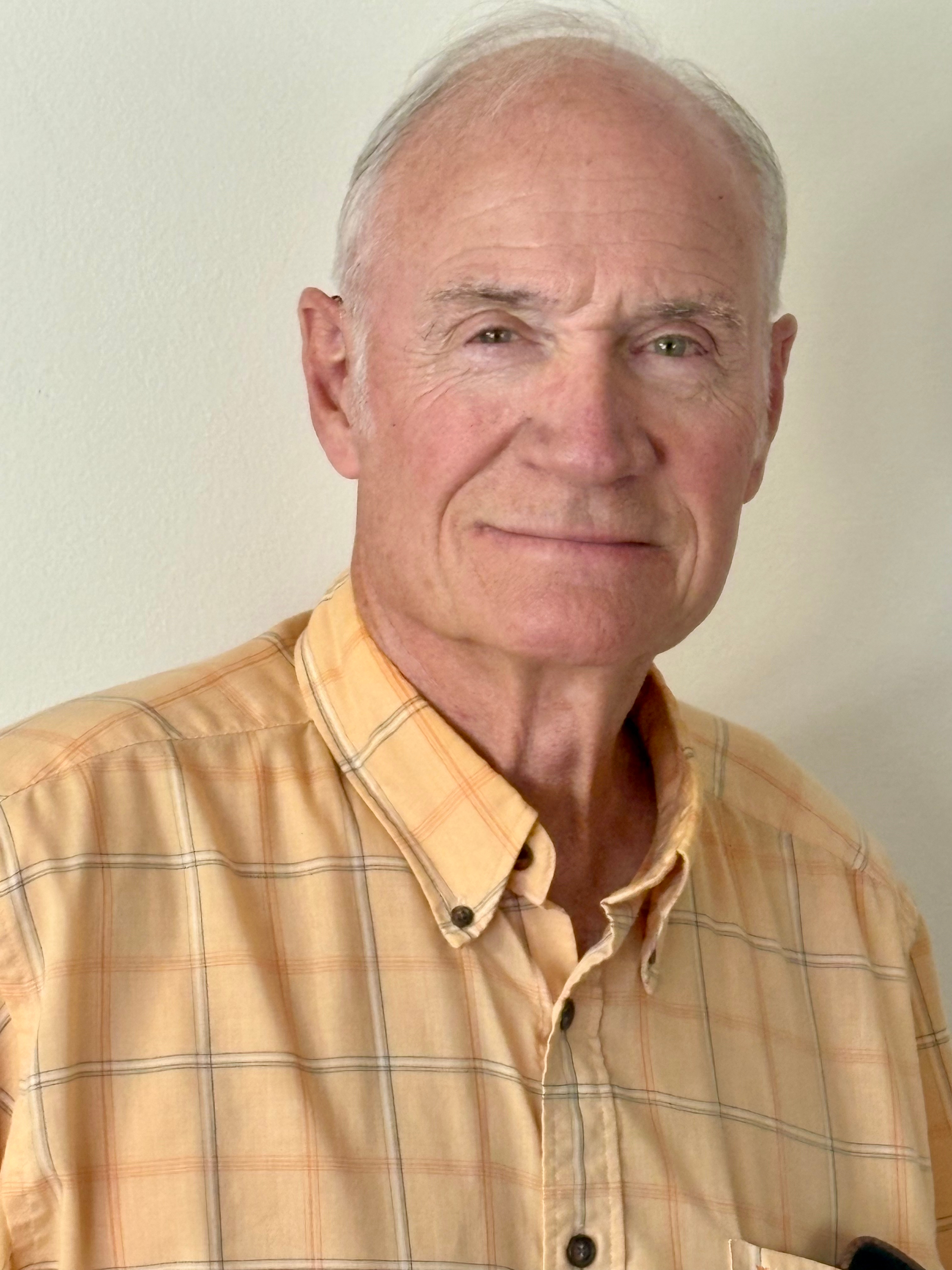 portrait of an elderly man with gray hair wearing a yellow plaid shirt, standing against a plain light-colored wall.
