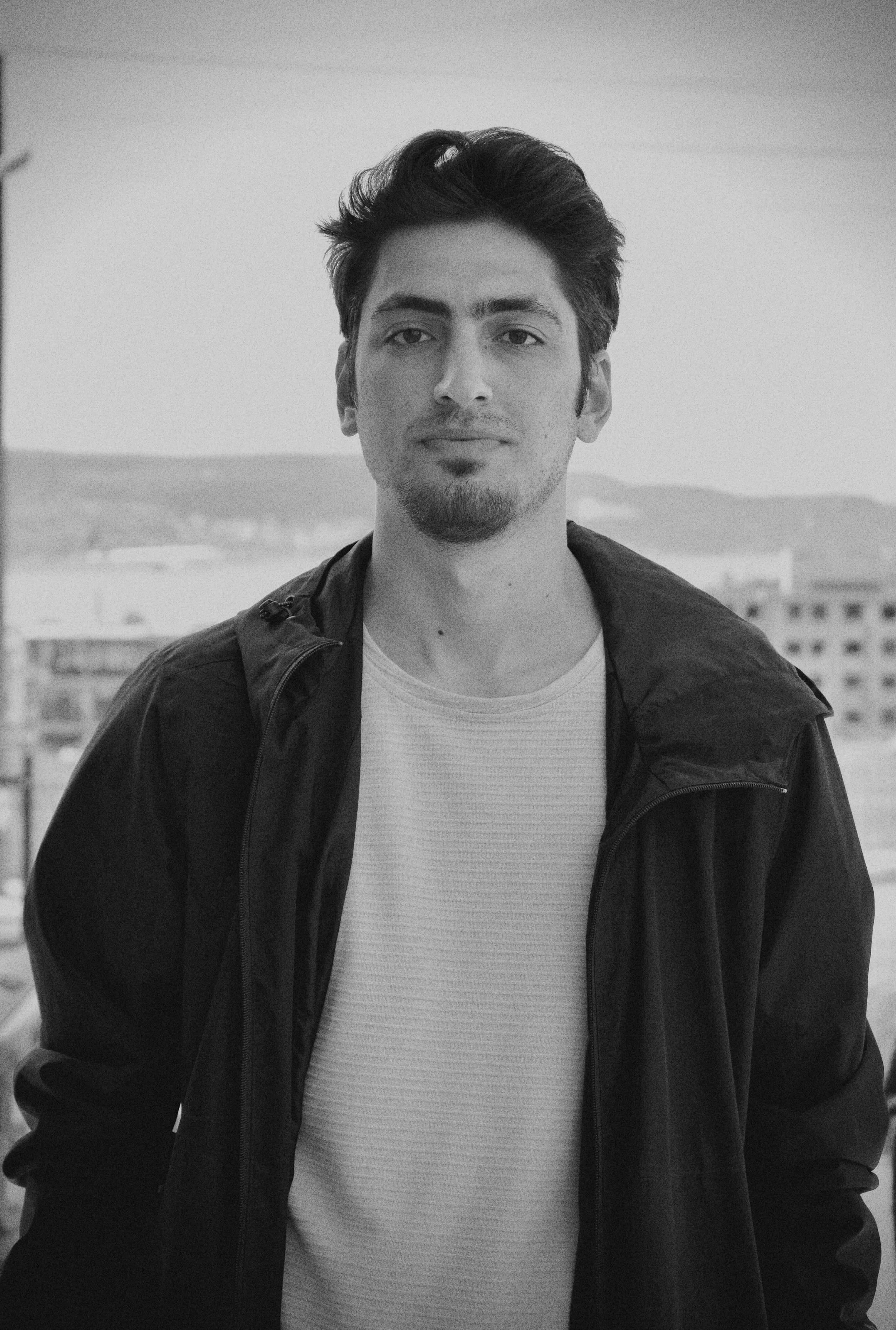 Black and white photo of a young man standing outdoors, wearing a jacket over a t-shirt, with buildings and hills in the background.