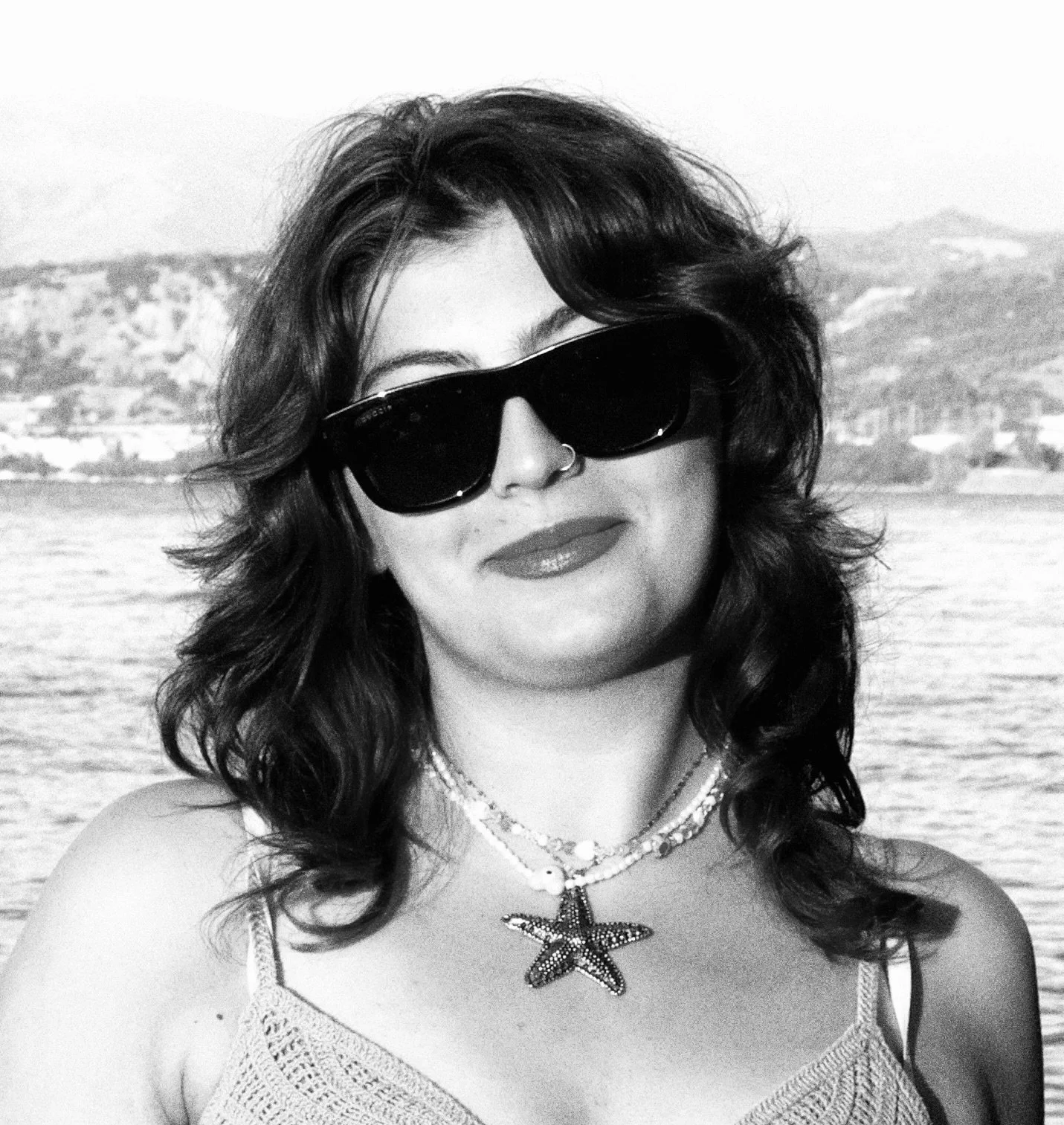 A woman with wavy hair wearing large sunglasses, a starfish necklace, and a tank top, standing near a body of water with hills in the background.