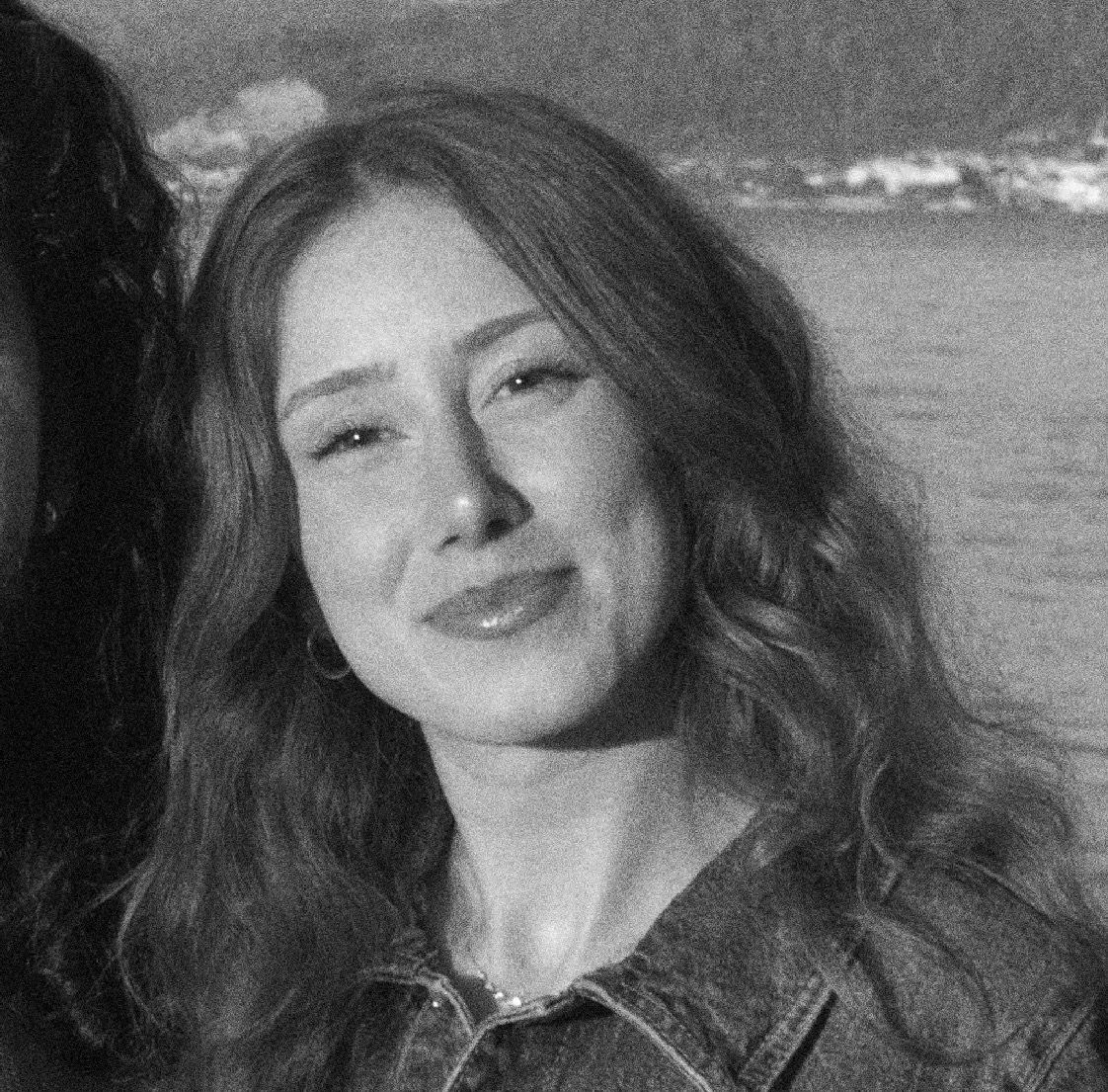 Black and white photo of a smiling woman with wavy hair, wearing a denim jacket, in front of a body of water.