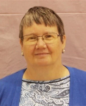 Middle-aged woman with short brown hair and glasses, wearing a blue cardigan over a patterned top, standing against a pink wall.