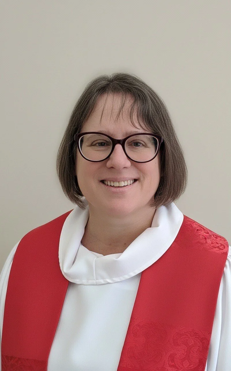 A woman dressed as a priest standing in front of a wooden altar with candles and flower arrangements, gesturing with her hands.
