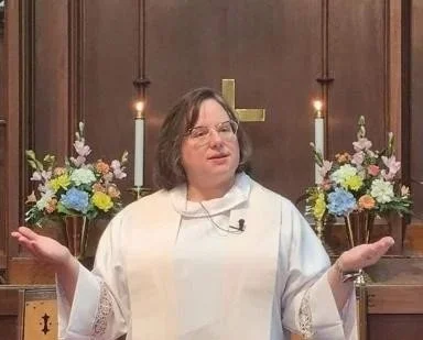 A woman dressed as a priest standing in front of a wooden altar with candles and flower arrangements, gesturing with her hands.