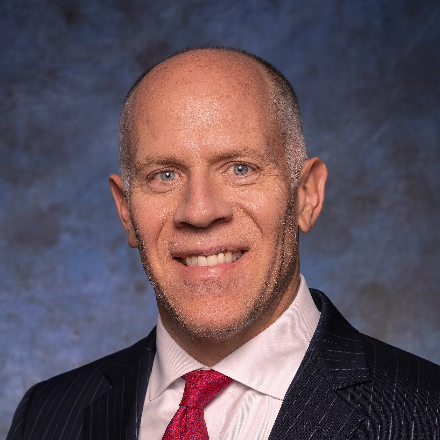 A professional headshot of a middle-aged man with light skin, blue eyes, a smile, short light hair, wearing a dark suit, pink shirt, and red tie, against a mottled blue background.