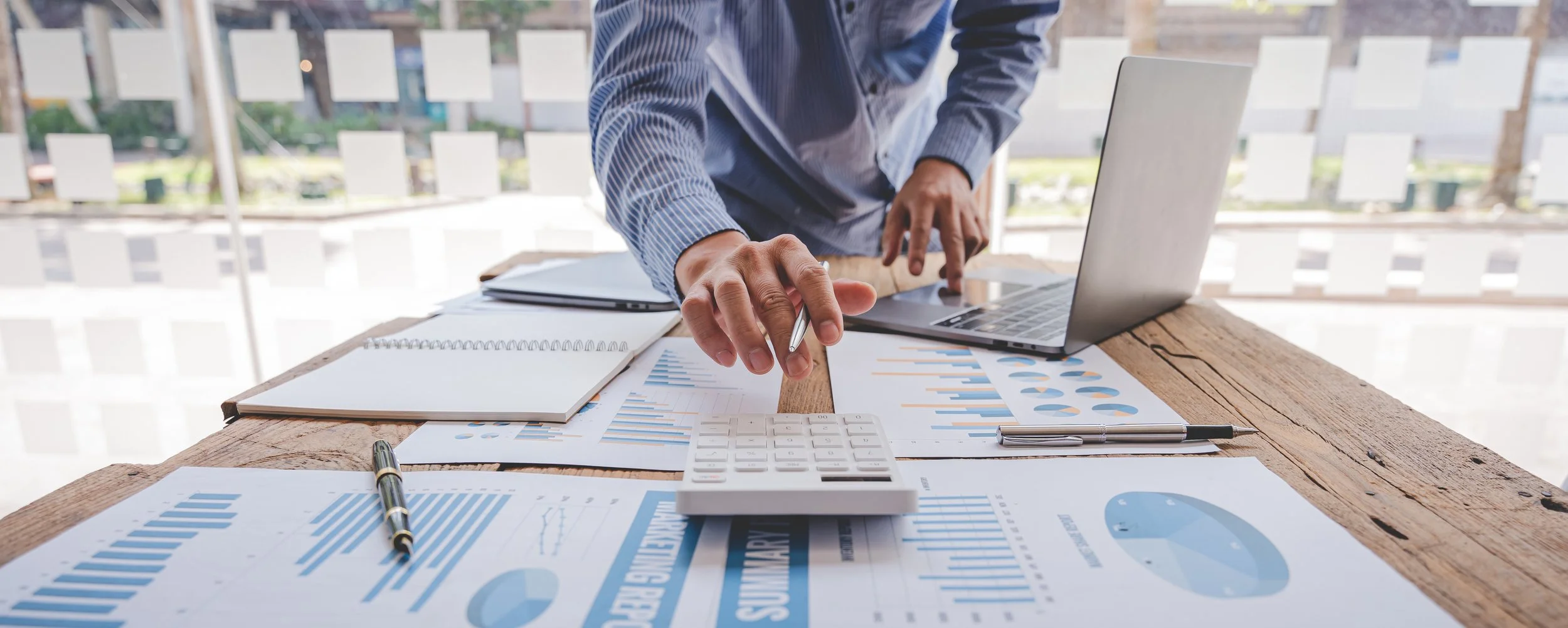 Person working at a desk with documents, a laptop, a calculator, notebooks, pens, and charts with graphs.