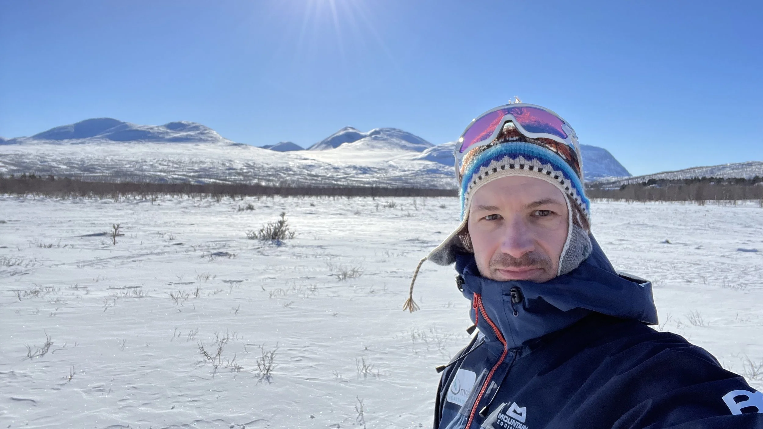 A man taking a selfie in a snowy landscape with mountains in the background and bright sunlight in the sky.