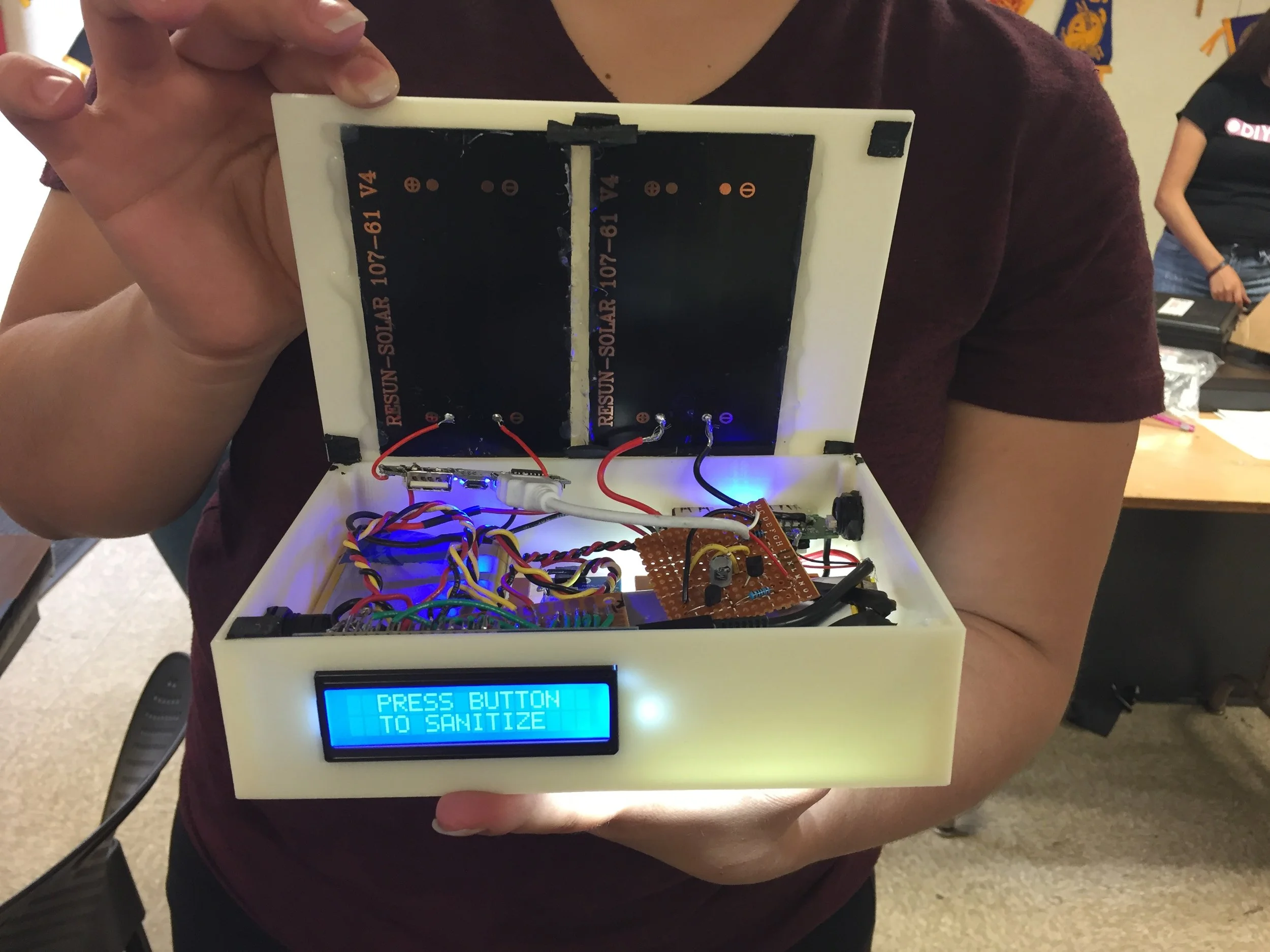 A person holding an open electronic device with a blue screen displaying the message 'PRESS BUTTON TO SANITIZE'. Inside, there are wires, circuit boards, and electronic components.