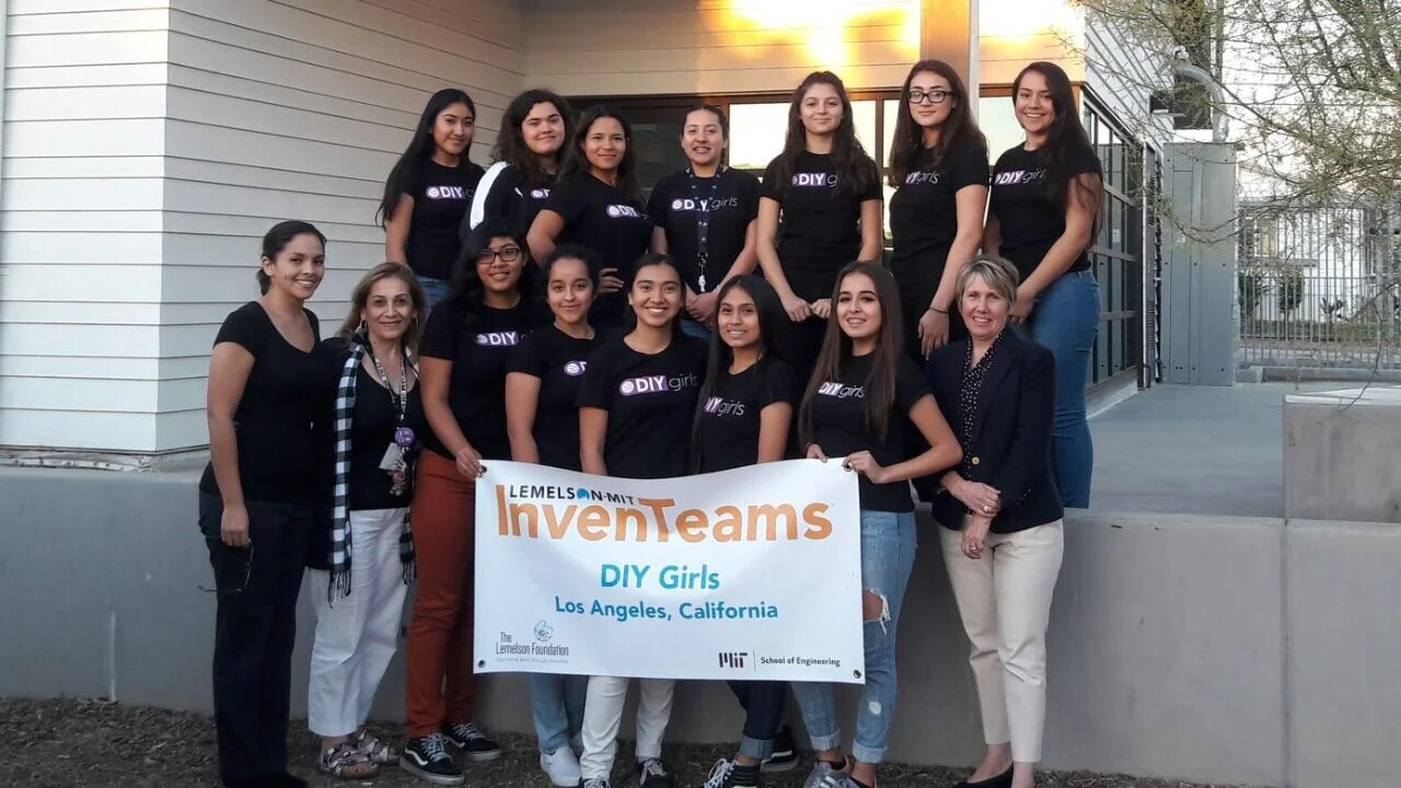 A group of young women and women teachers posing outdoors on steps, holding a banner that reads 'LEMELSON-MIT InvenTeams DIY Girls Los Angeles, California'.